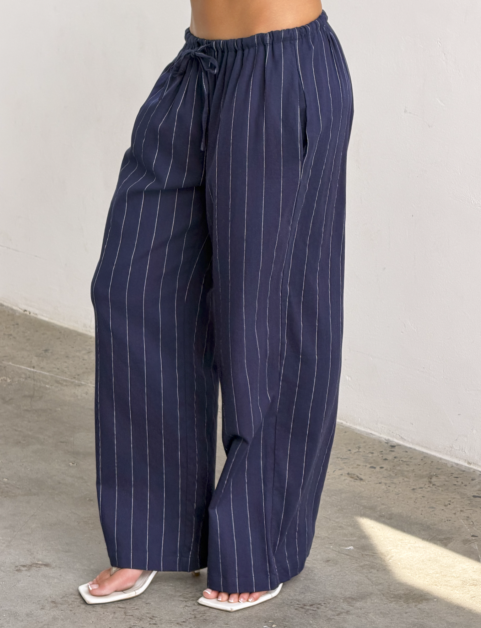 Someone wearing the TIGERMIST LAINEY PANT in navy pinstripe—loose-fitting linen pants with an elastic waist—paired with white sandals, stands on a concrete floor against a light-colored wall.