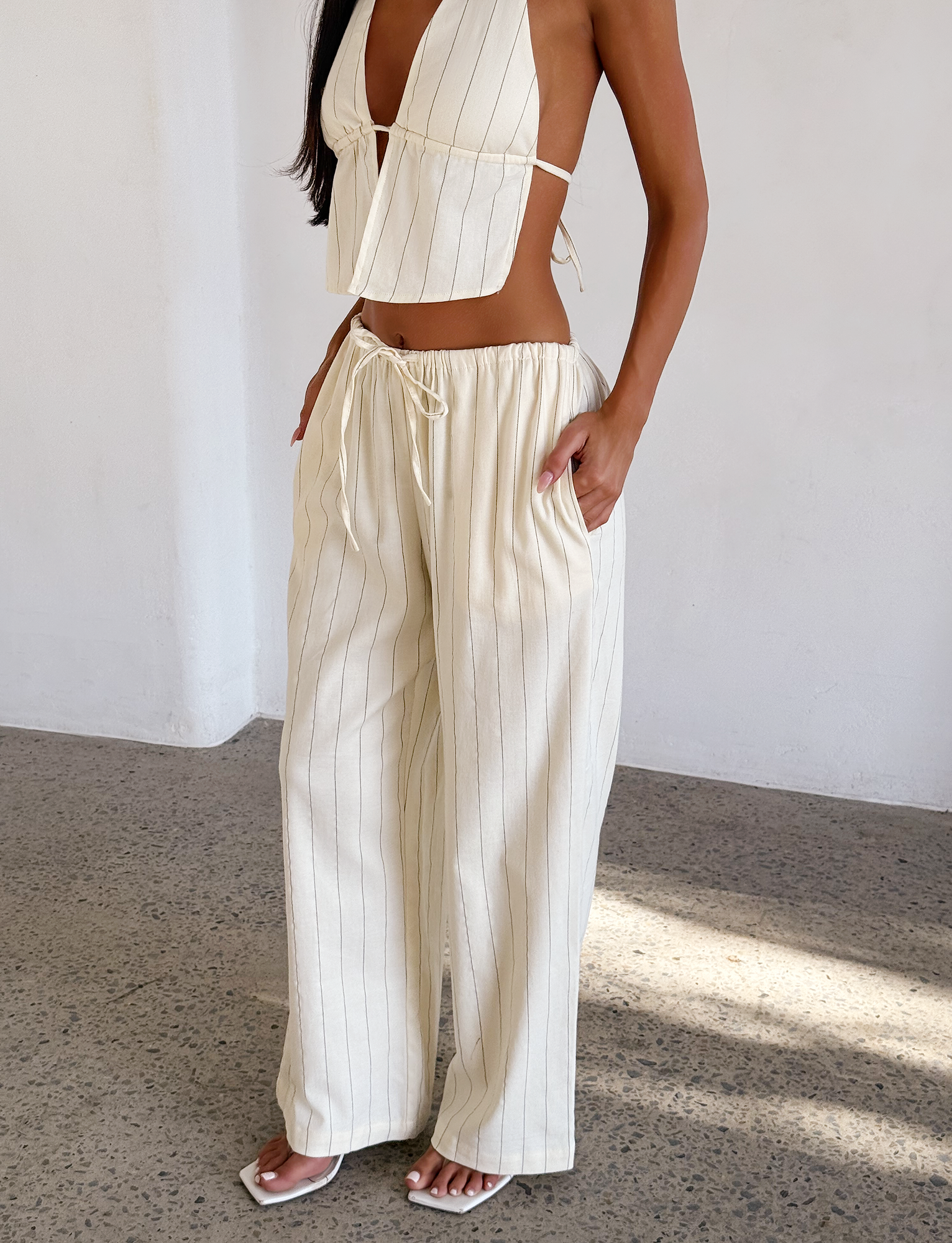 Woman wears the TIGERMIST LAINEY PANT - BEIGE STRIPE, a wide-leg linen style paired with a halter crop top and white sandals, standing indoors on a speckled floor.