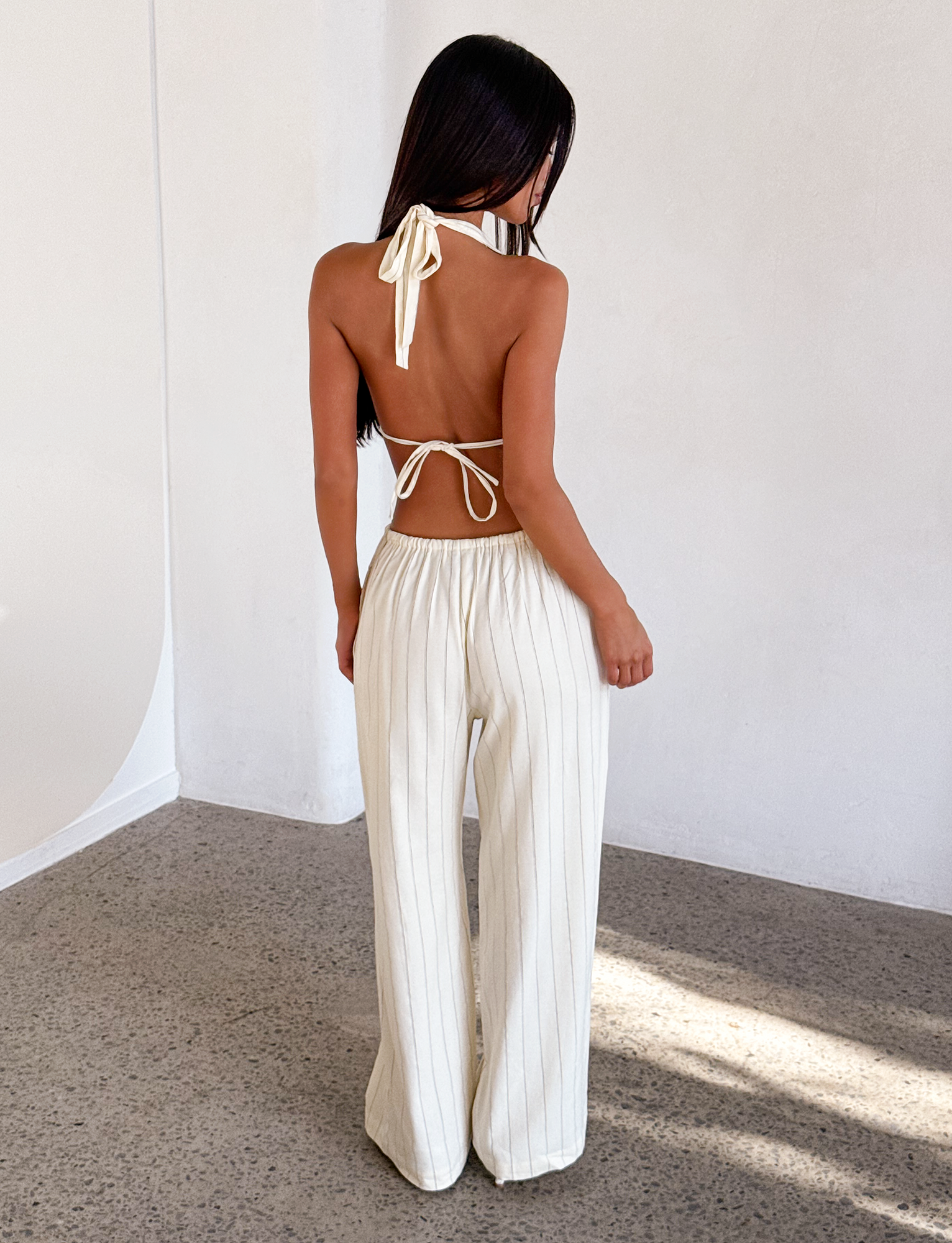 A woman stands indoors with her back to the camera, wearing the TIGERMIST LAINEY PANT in beige stripe—a pair of wide leg, high-waisted linen pants. The room has neutral walls and a speckled floor.