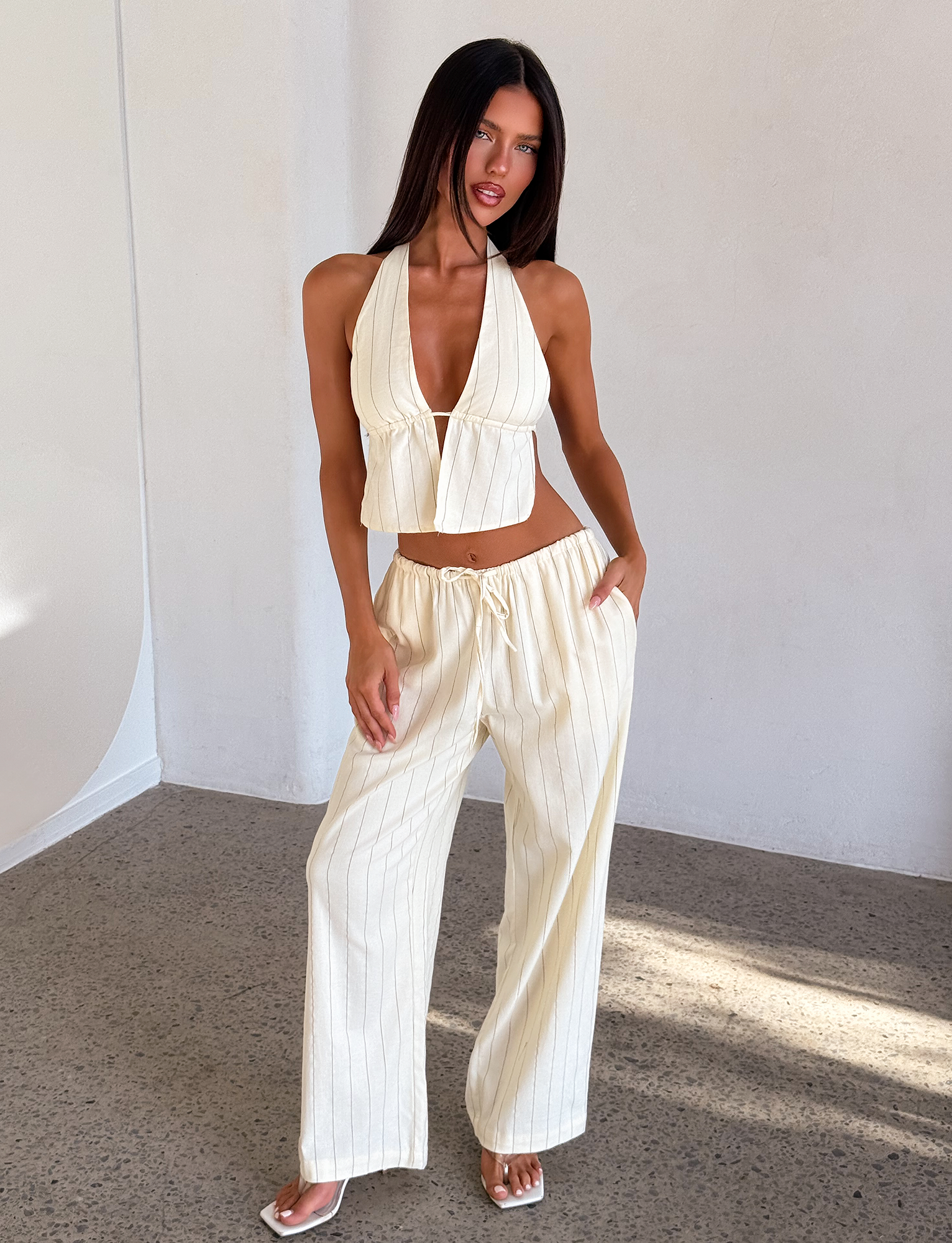 A woman stands indoors wearing TIGERMIST's LAINEY PANT in beige stripe, paired with a matching pinstripe halter top and white sandals. She poses with one hand in her pocket as sunlight streams in.