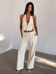 A woman stands indoors wearing the cream-colored, striped CL - LAINEY PANT by Tiger Mist USA with a matching halter top. She poses confidently with hands in her pockets, looking forward.