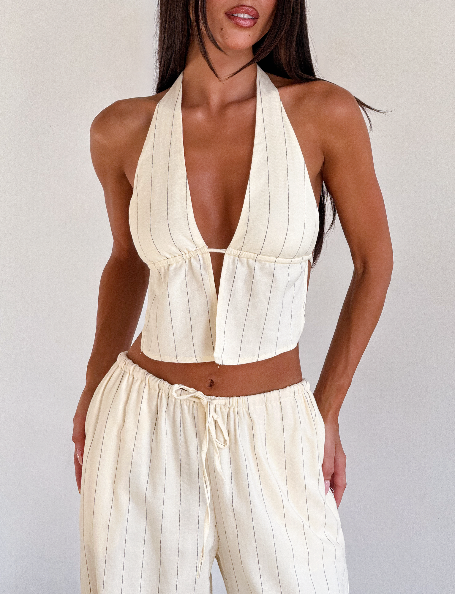 A woman wears the TIGERMIST LAINEY TOP in beige stripe, an open back linen halter with adjustable ties, paired with matching drawstring pants, standing against a plain light background.