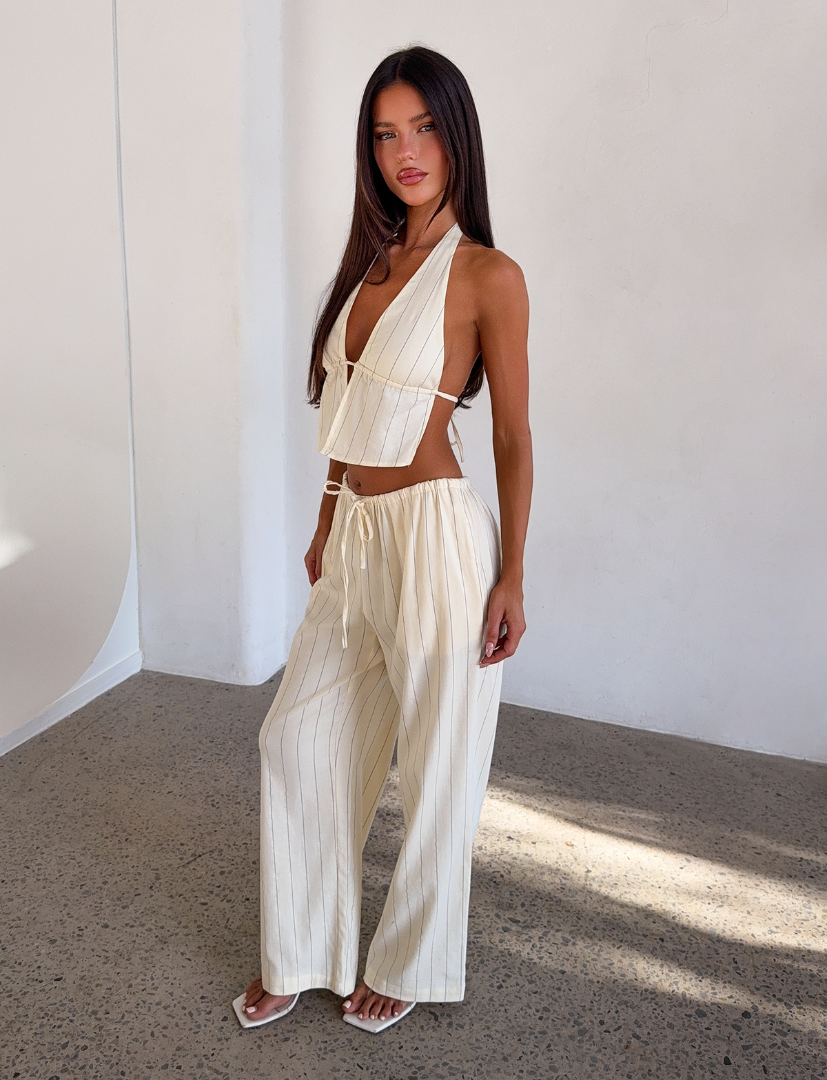 A woman poses indoors against a white wall wearing the TIGERMIST LAINEY PANT in beige stripe, styled with a cream backless halter crop top, white sandals, and sporting long dark hair.