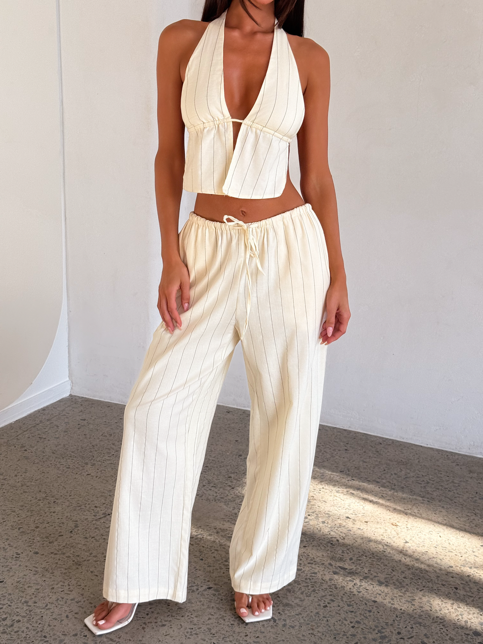 A woman wears the TIGERMIST LAINEY PANT - BEIGE STRIPE, featuring wide legs and a drawstring waist, paired with a matching pinstripe halter crop top and white open-toe heels. She stands indoors against a neutral wall.