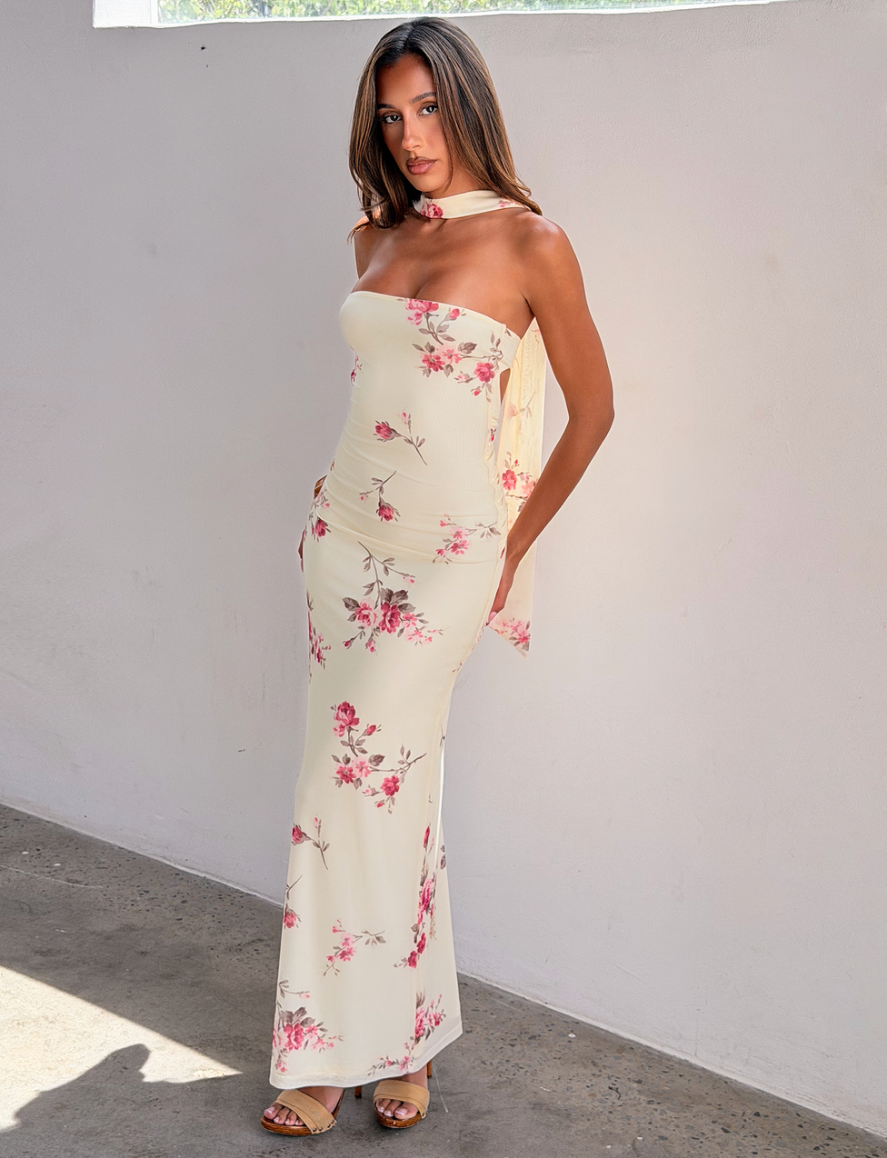 A woman with straight brown hair and a neutral expression stands by a light wall wearing the TIGERMIST DANIELA SCARF DRESS - YELLOW FLORAL, a strapless, floor-length gown with matching scarf and beige sandals.