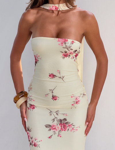 A woman wears the TIGERMIST DANIELA SCARF DRESS - YELLOW FLORAL, a strapless, fitted dress with pink floral patterns. She accessorizes with a matching scarf and chunky bracelets on her left wrist against a plain white background.