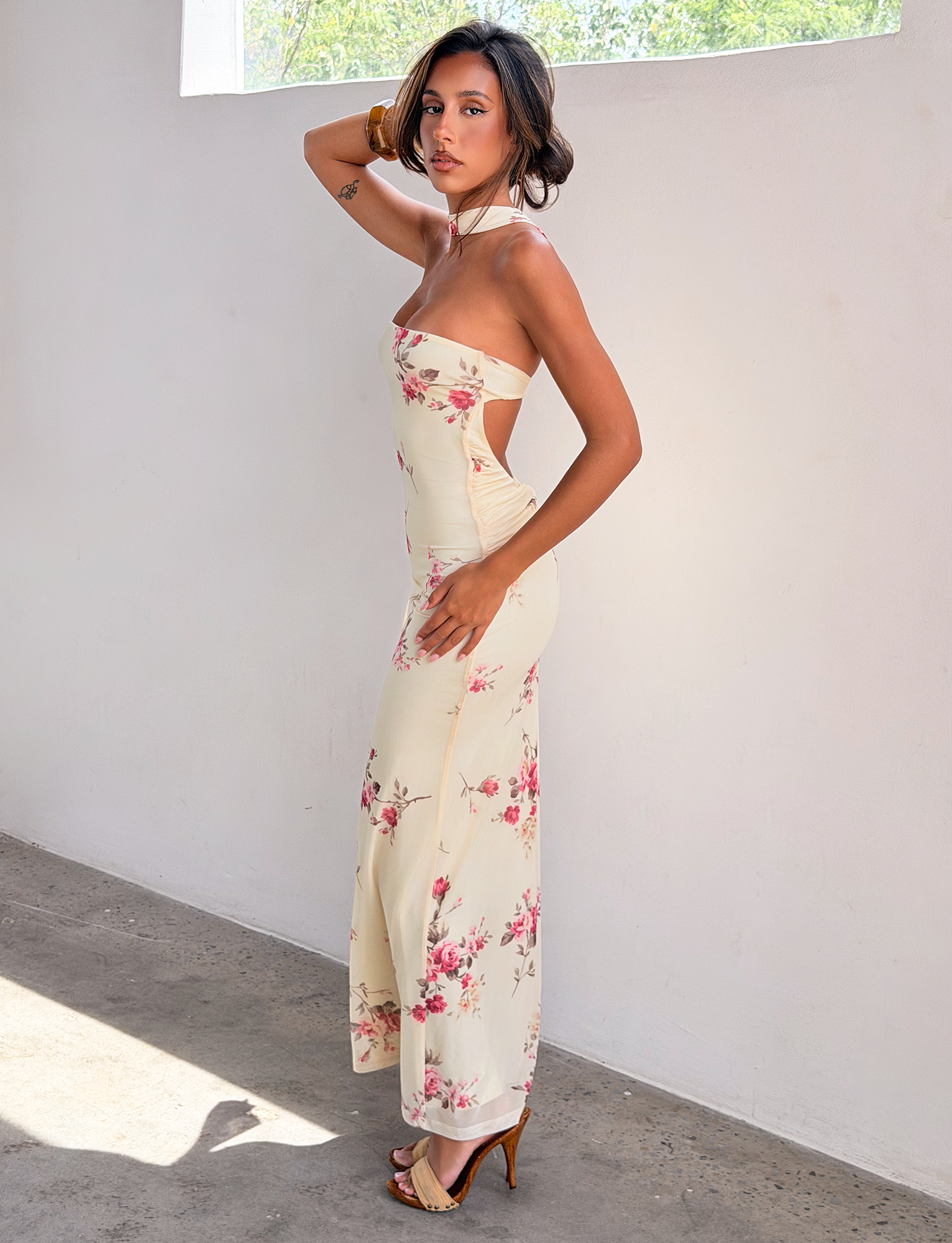 A woman stands indoors against a white wall, modeling the TIGERMIST DANIELA SCARF DRESS - YELLOW FLORAL. She poses sideways, one hand on her head, as sunlight pours in from above.