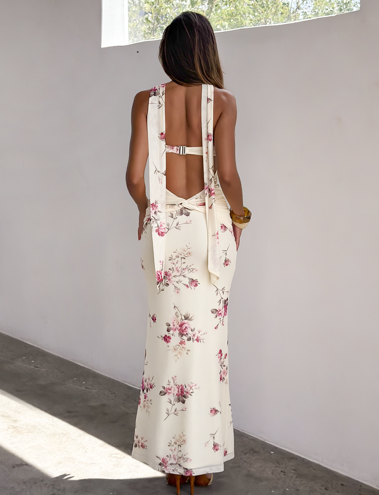A woman stands indoors, sunlight streaming in as she faces a white wall, elegantly wearing the TIGERMIST DANIELA SCARF DRESS in yellow floral with open back, tie, and buckle details.