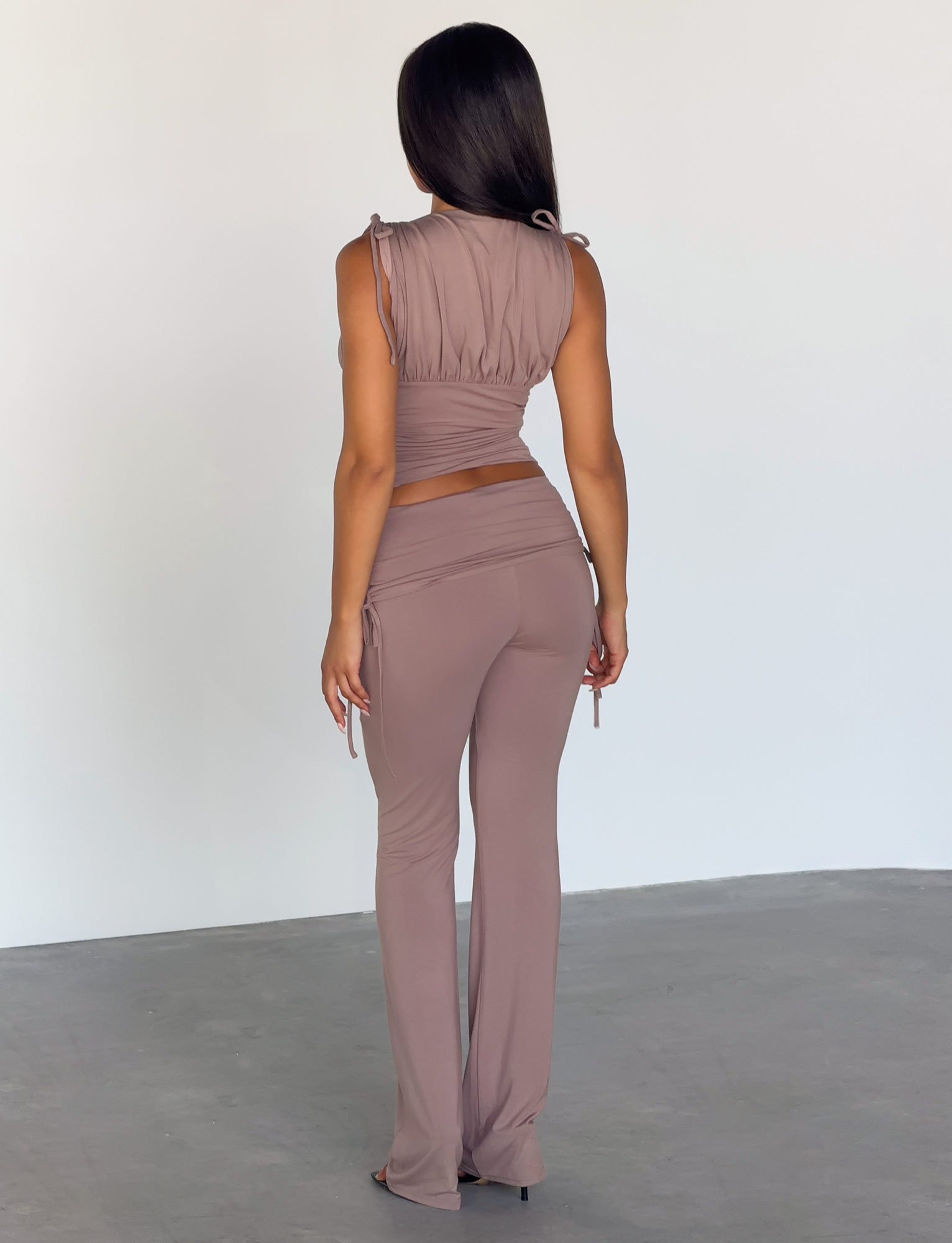 A woman with straight dark hair stands indoors, wearing the HALONA PANT - MOCHA by TIGERMIST. The mocha pants have a subtle flared silhouette and are paired with a sleeveless ruched mauve top. She faces away in a white-walled, concrete-floored space.