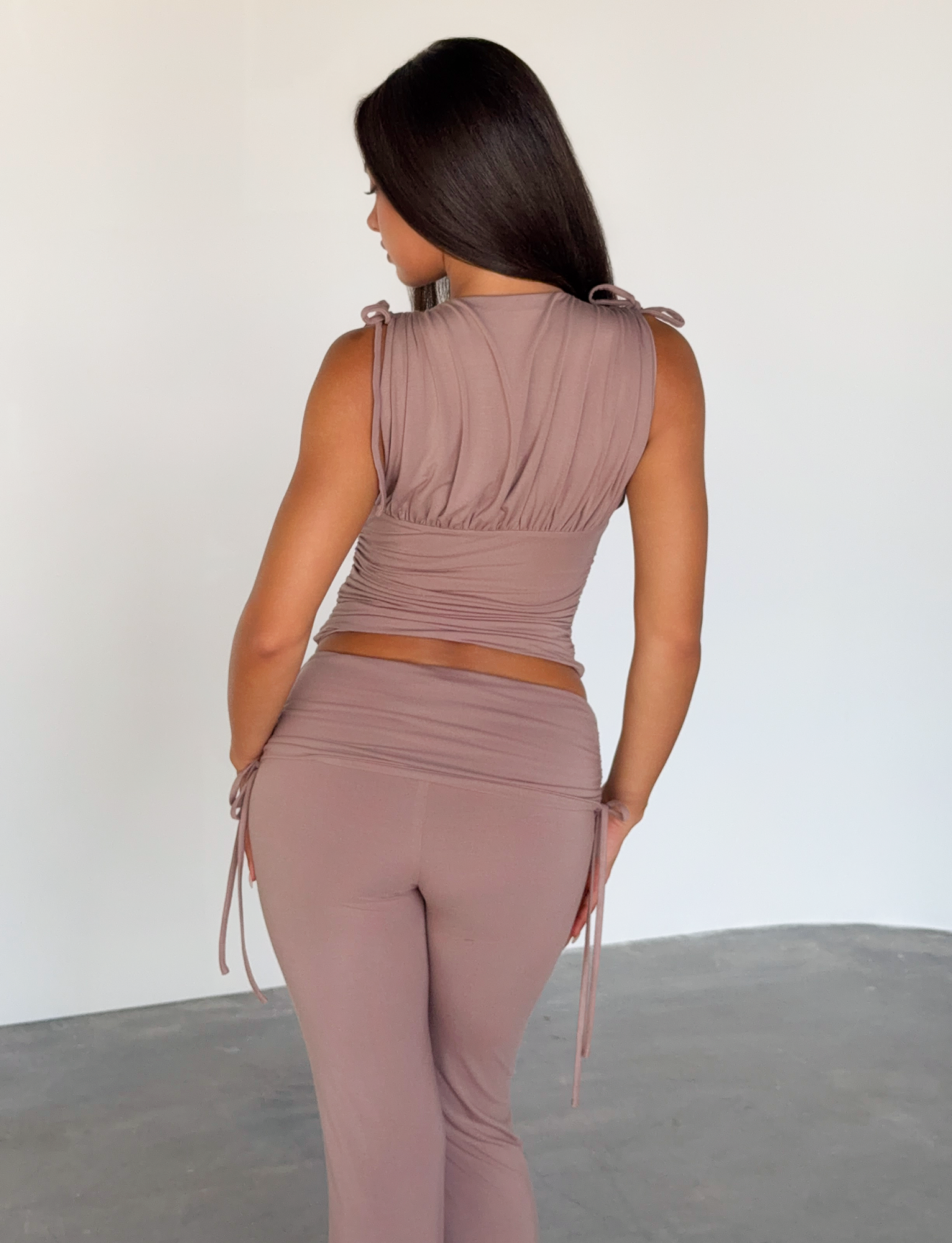 A woman with long dark hair stands with her back to the camera, wearing the TIGERMIST HALONA TOP in mocha—a sleeveless, ruched bodice top with adjustable shoulder ties—in a minimalist indoor setting.