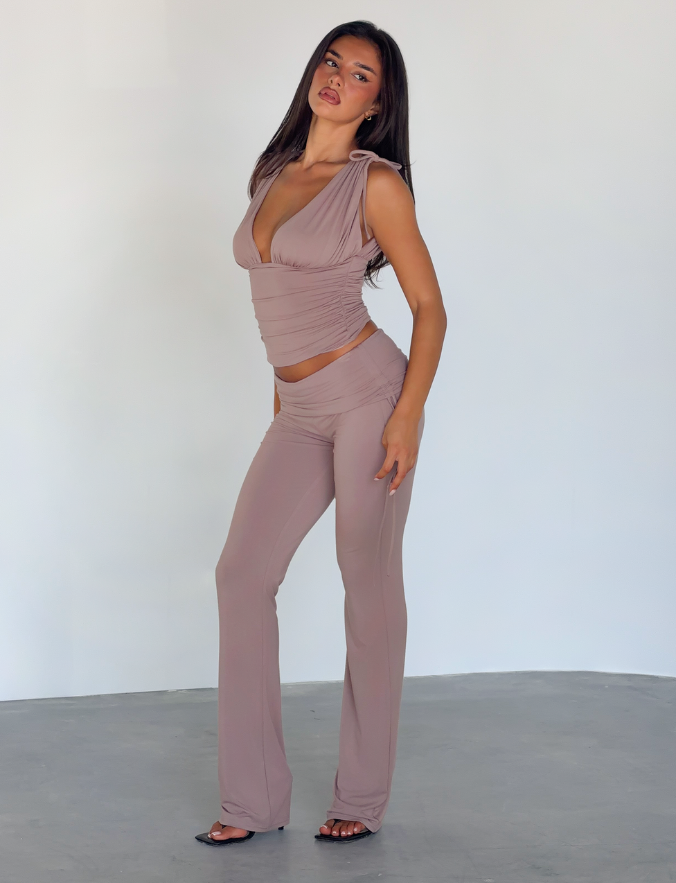 A woman with long dark hair stands confidently against a plain background, wearing TIGERMIST's HALONA PANT in mocha—a fitted, flared silhouette—paired with a matching sleeveless top and open-toed heels.