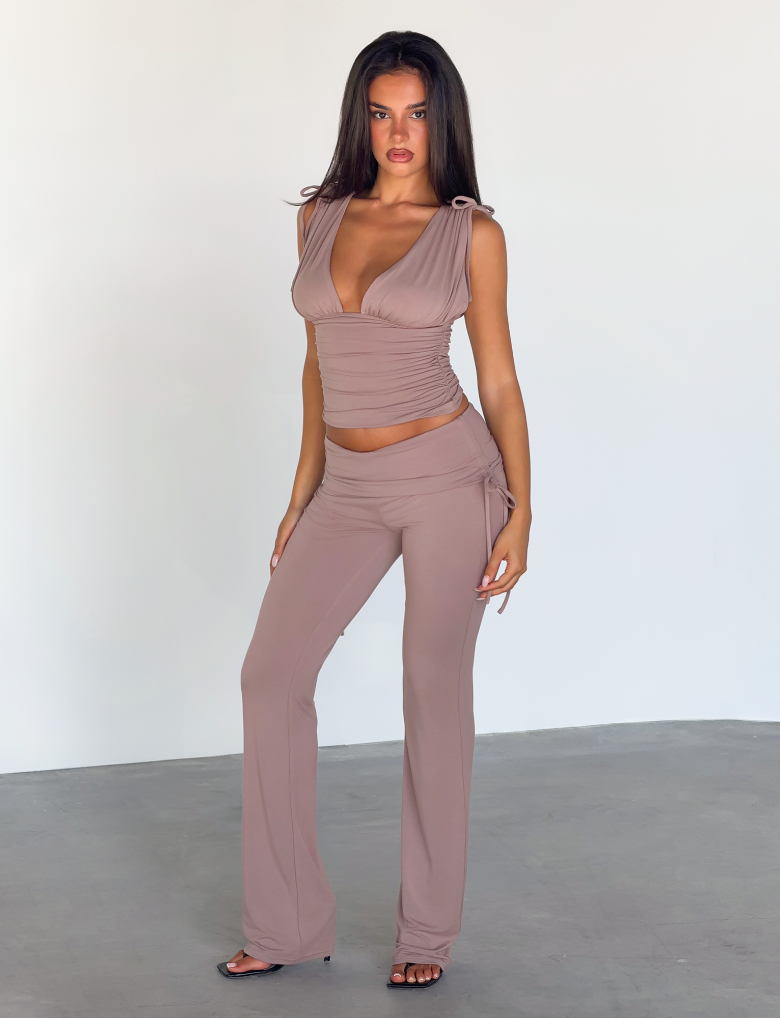A woman with long dark hair poses confidently indoors against a white wall, wearing the TIGERMIST HALONA TOP in mocha—a sleeveless, deep V-neck ruched bodice—paired with flared pants.
