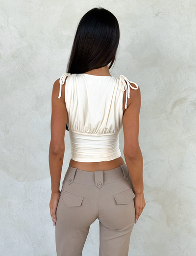 A woman with long dark hair faces away from the camera, wearing the TIGERMIST HALONA TOP in cream with a plunging ruched bodice and adjustable shoulder ties, paired with high-waisted beige pants. She stands before a light textured wall.