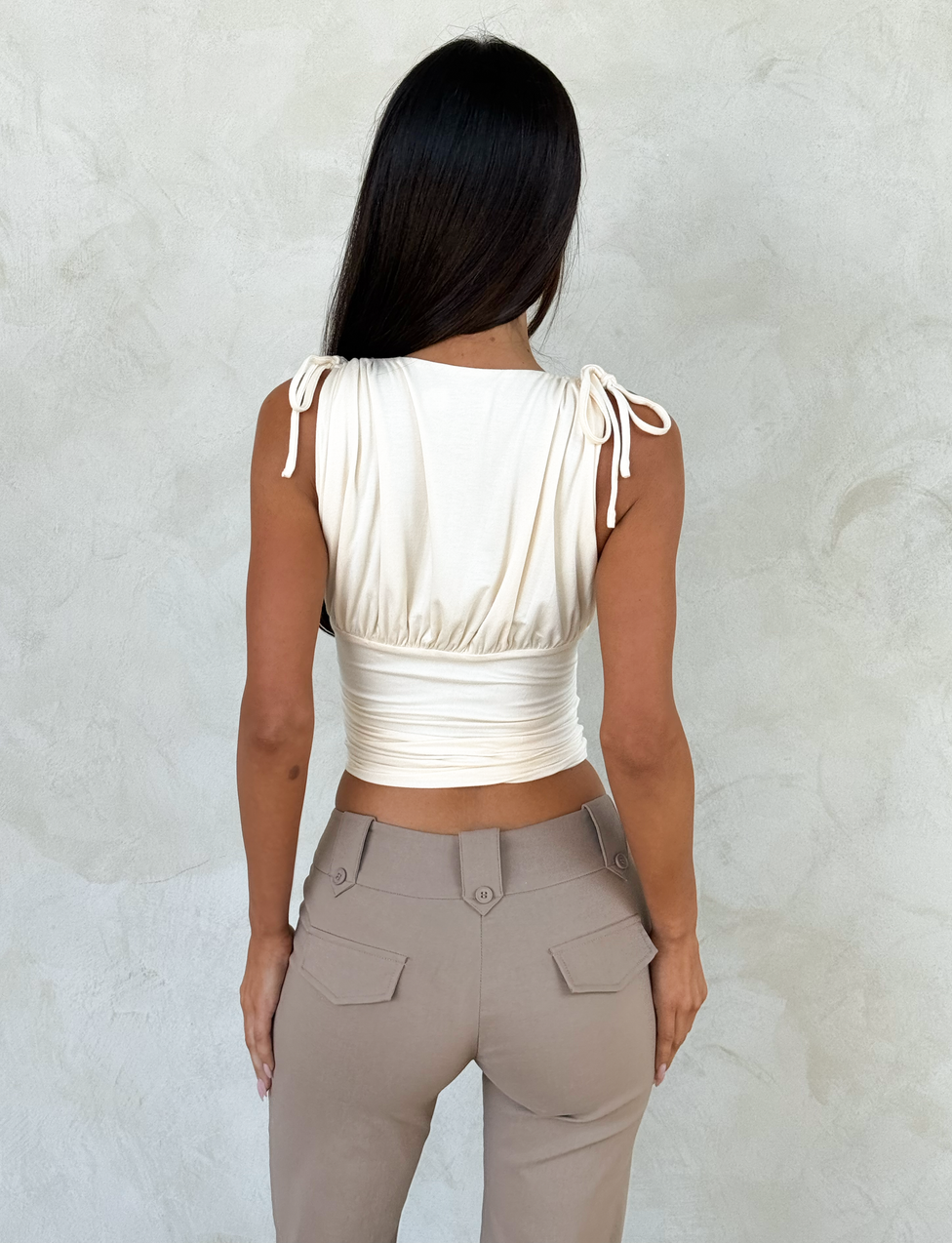 A woman with long dark hair faces away from the camera, wearing the TIGERMIST HALONA TOP in cream with a plunging ruched bodice and adjustable shoulder ties, paired with high-waisted beige pants. She stands before a light textured wall.