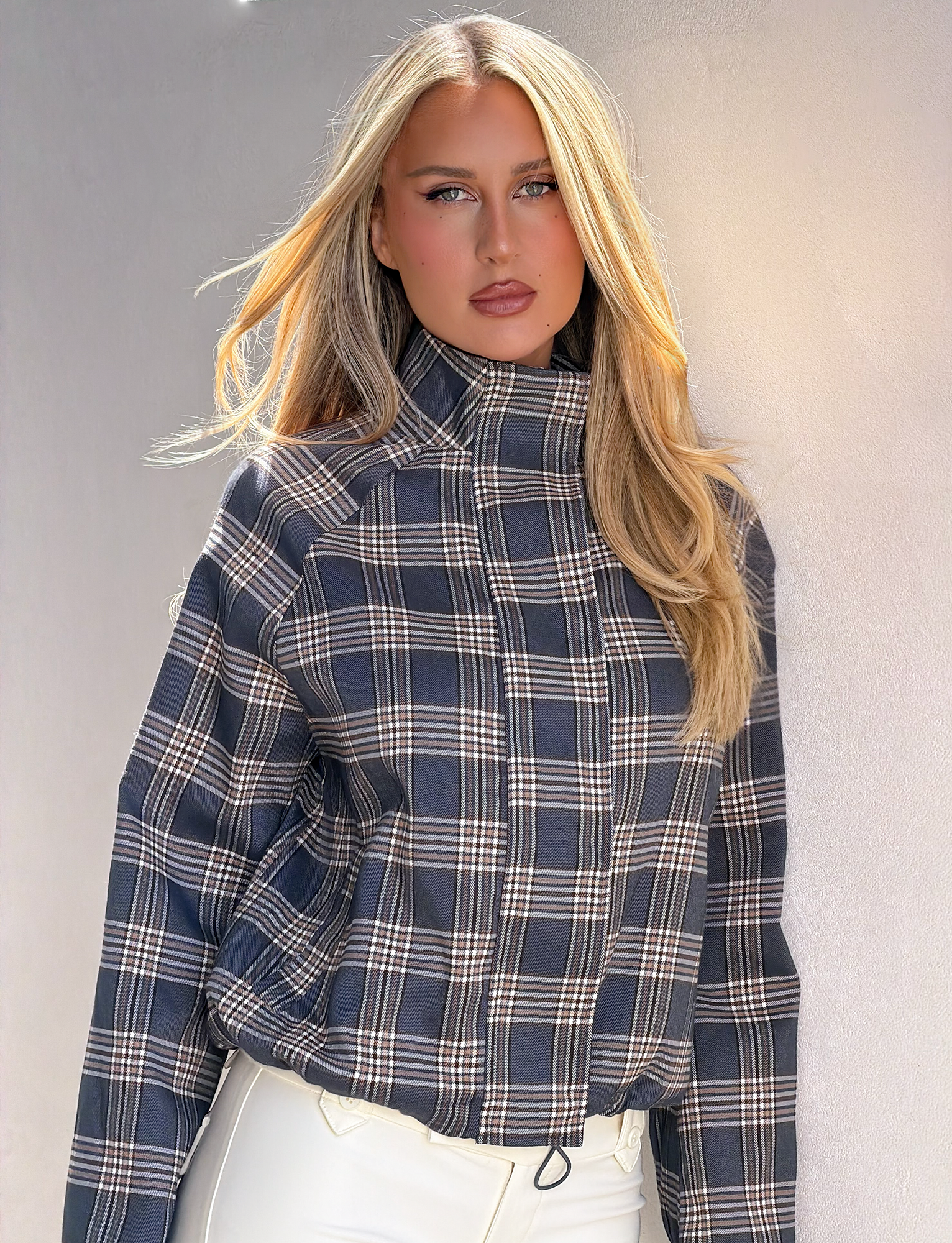 A woman with long blonde hair wears the TIGERMIST LENNEX JACKET in grey check, styled with white pants and standing against a light-colored wall.