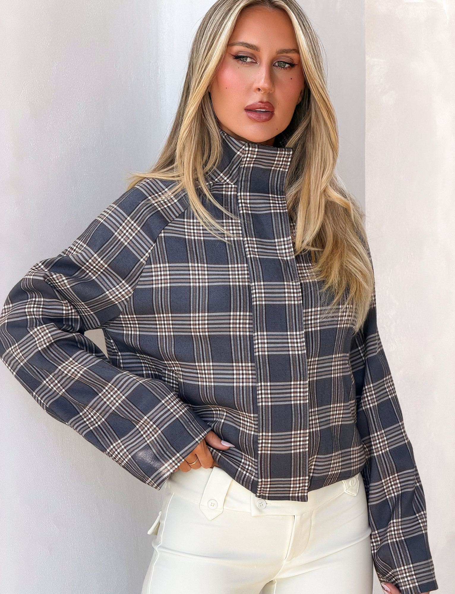 A woman with long blonde hair wears the TIGERMIST LENNEX JACKET in grey check, featuring an oversized fit and mock neck collar, paired with white pants and standing against a white wall.