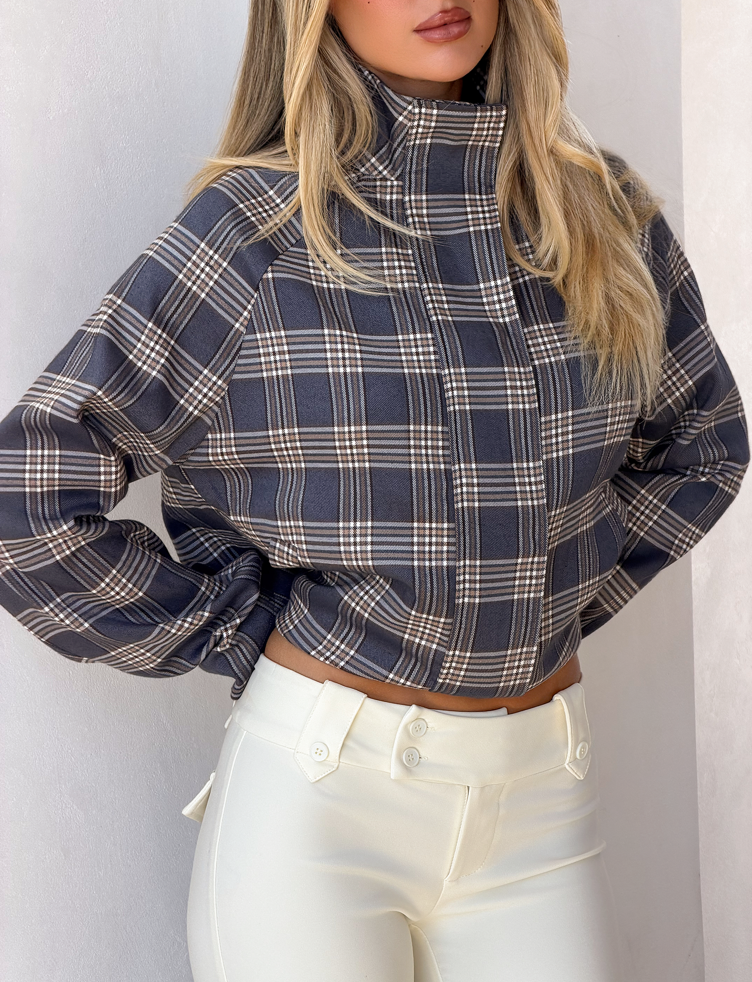 A woman wearing the TIGERMIST LENNEX JACKET in grey check stands against a light background. She pairs it with high-waisted white pants, her blonde hair loose and face partially visible.
