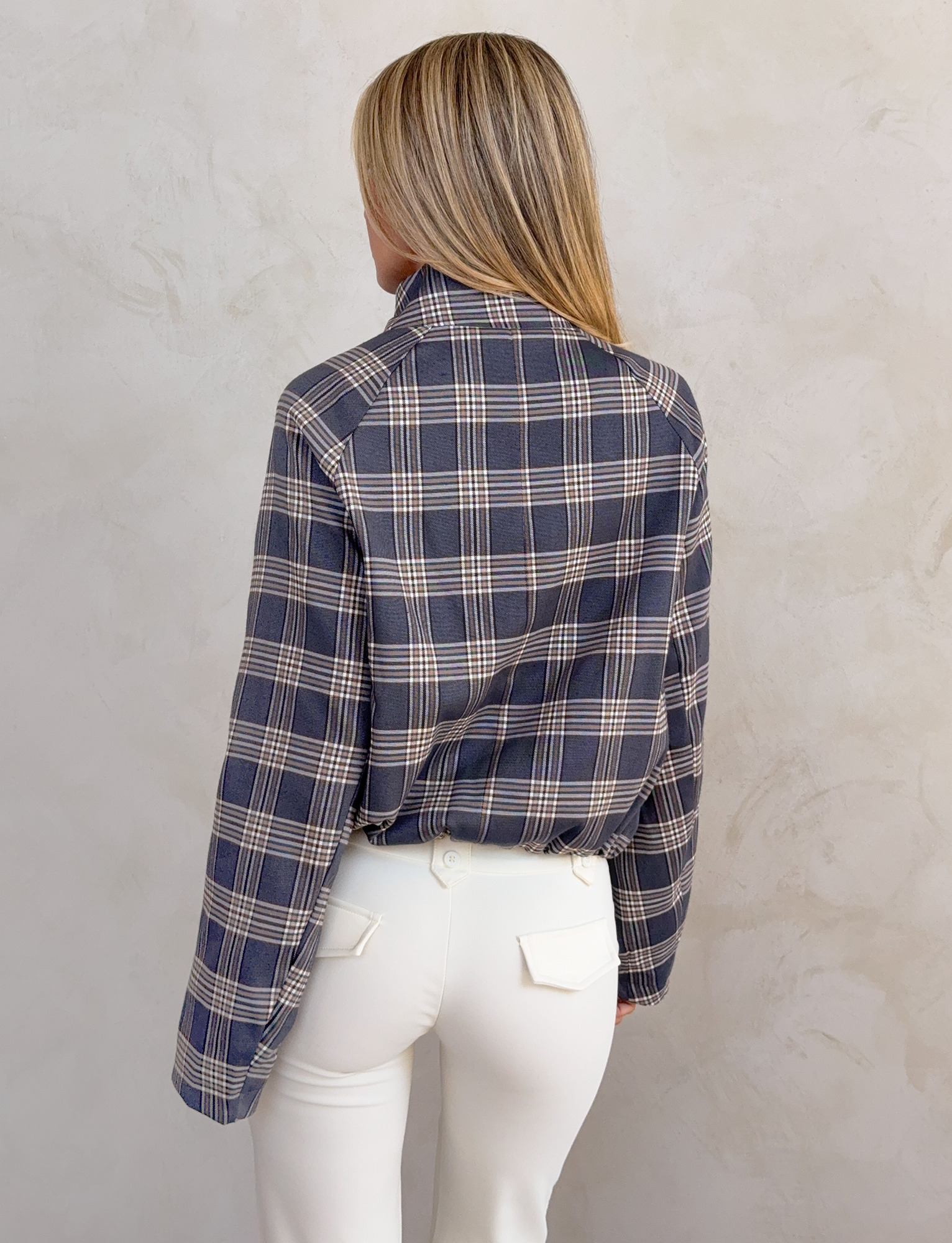 Seen from behind, a person with long blonde hair wears the TIGERMIST LENNEX JACKET in grey check, featuring a bungee drawstring hem, paired with white pants against a light textured wall.