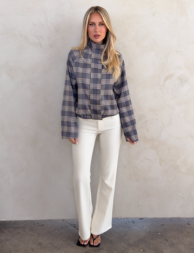 A woman with long blonde hair and a neutral expression stands against a light wall wearing the CL - LENNEX JACKET by Tiger Mist USA, styled with white high-waisted pants and black open-toe sandals.