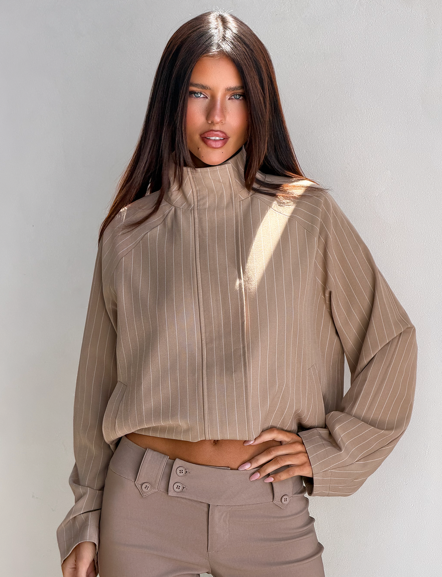 A woman with straight brown hair poses against a light-colored wall wearing the TIGERMIST LENNEX JACKET in tan pinstripe, exuding effortless style with one hand on her hip.