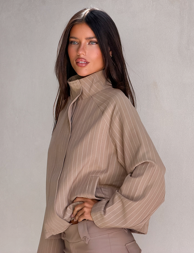 A woman with long dark hair stands against a light wall, confidently posing in the TIGERMIST LENNEX JACKET - TAN PINSTRIPE.