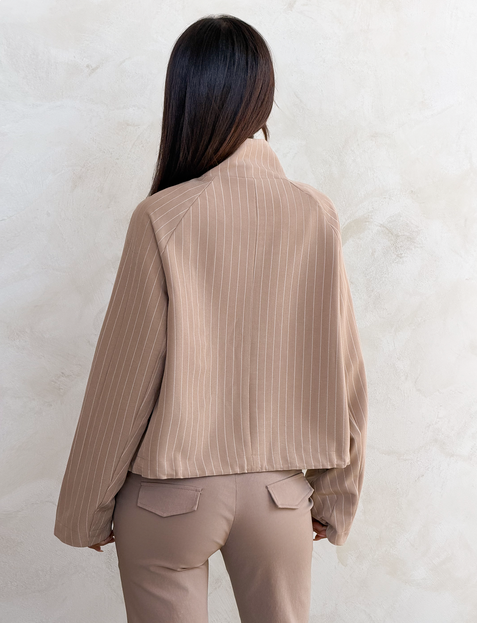 A woman with long dark hair stands with her back to the camera, wearing the TIGERMIST LENNEX JACKET in tan pinstripe, paired with matching pants, against a light textured wall.