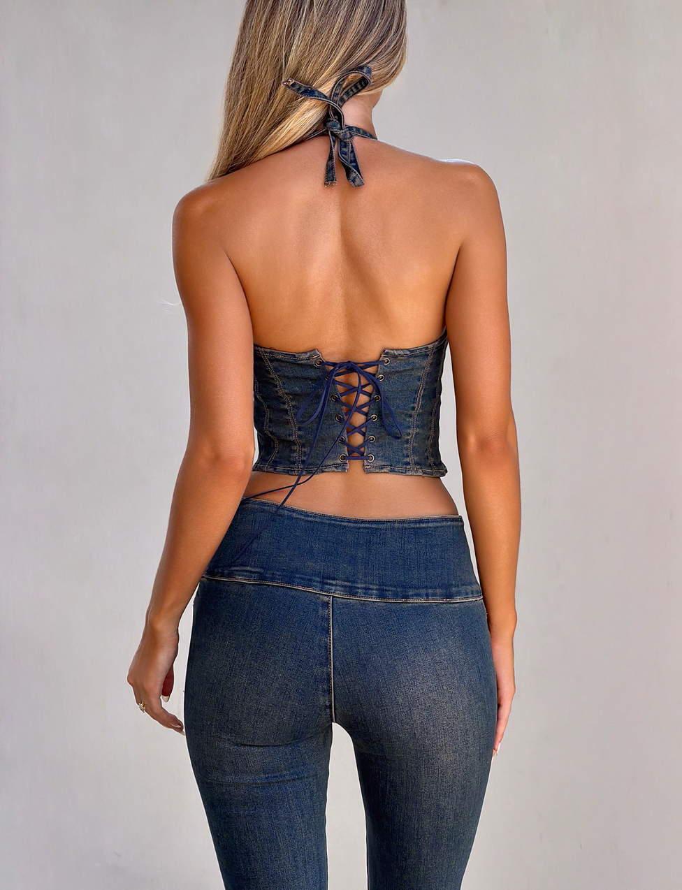 A woman faces away, wearing the TIGERMIST RIONA TOP - MIDWASH, a dark denim corset halter with lace-up back, paired with matching high-waisted skinny jeans against a plain light background.