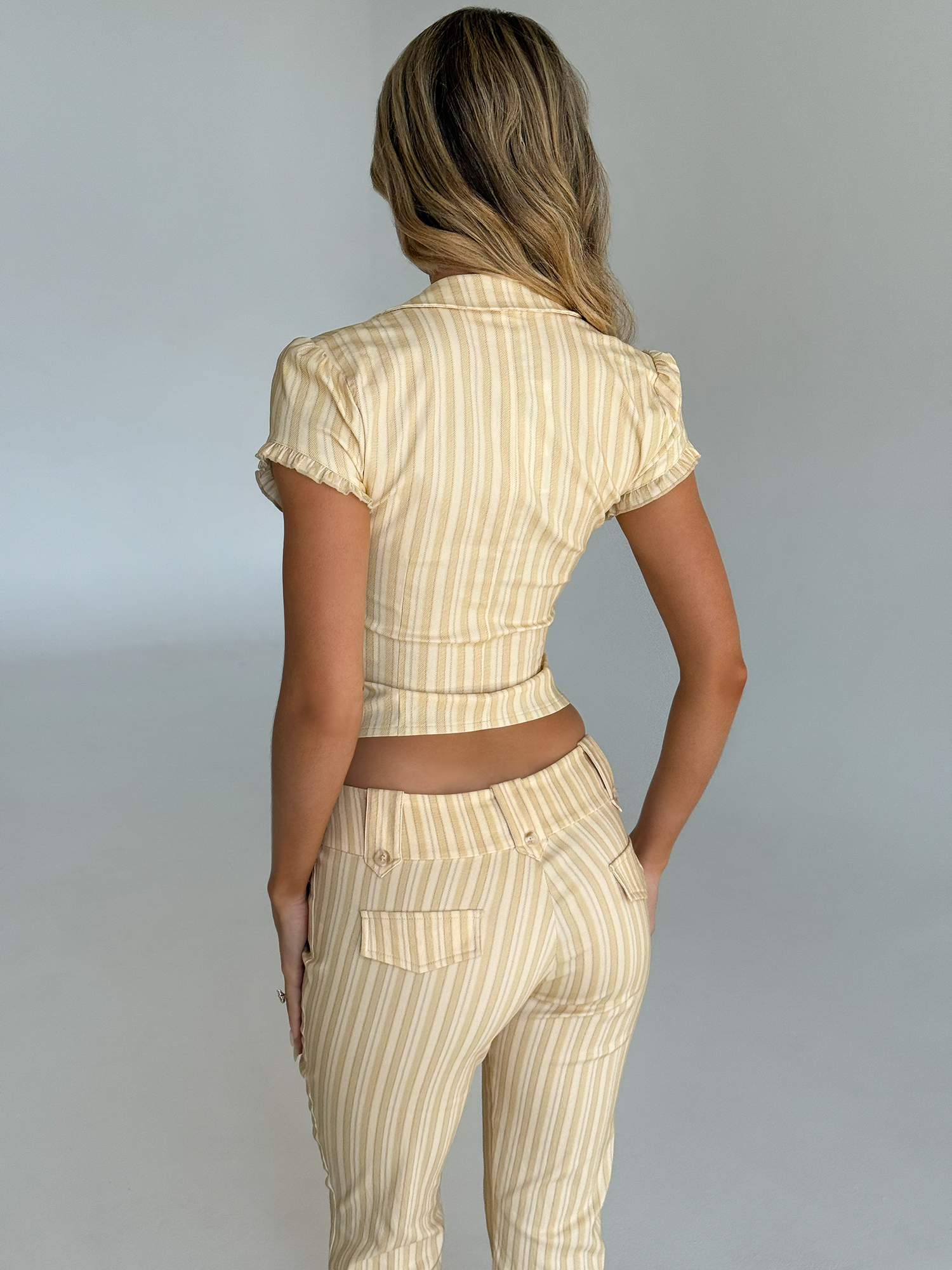 A woman with wavy blonde hair faces away, wearing the TIGERMIST KITTIE CAPRI yellow stripe fitted, short-sleeved top and matching high-waisted pants in a minimalist, light-toned room.