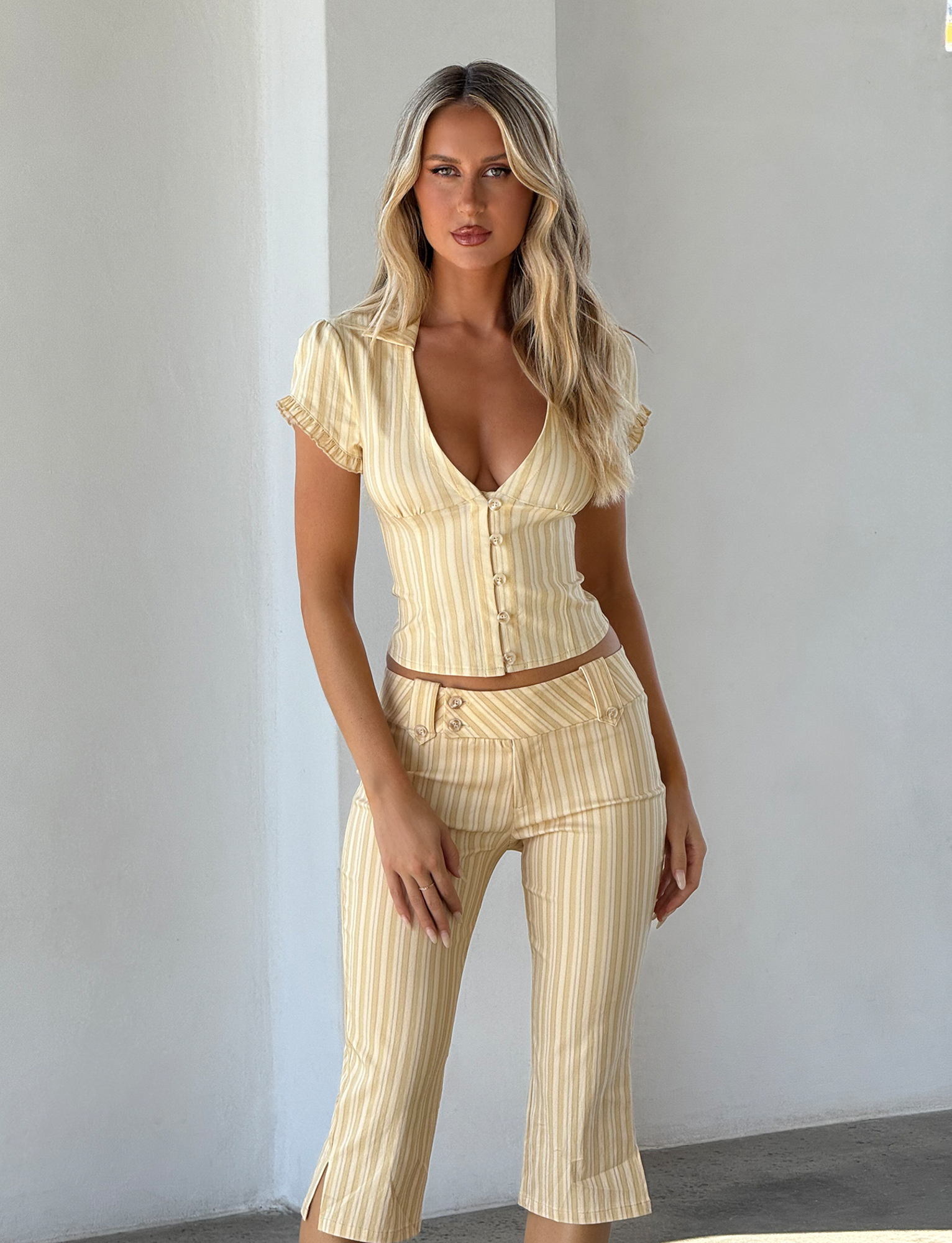 A woman with long blonde hair stands against a white wall, wearing the TIGERMIST KITTIE SHIRT in yellow stripe, made from stretch suiting fabric, paired with matching cropped pants. She faces forward with a neutral expression.