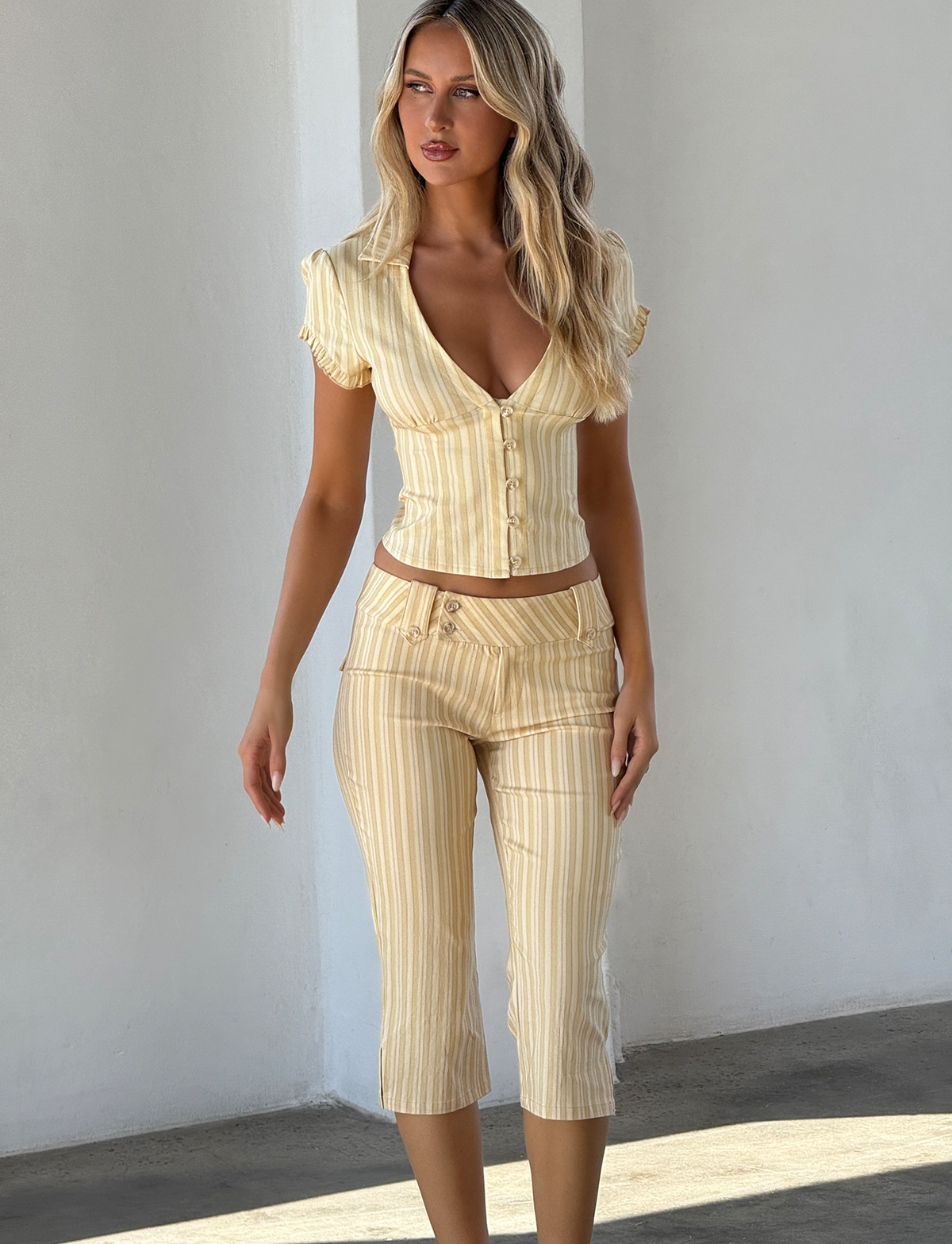 A woman with long blonde hair wears TIGERMIST's KITTIE CAPRI - YELLOW STRIPE: a light yellow, short-sleeve deep V-neck buttoned top and matching high-waisted cropped pants in stretch suiting fabric, standing against a white wall.