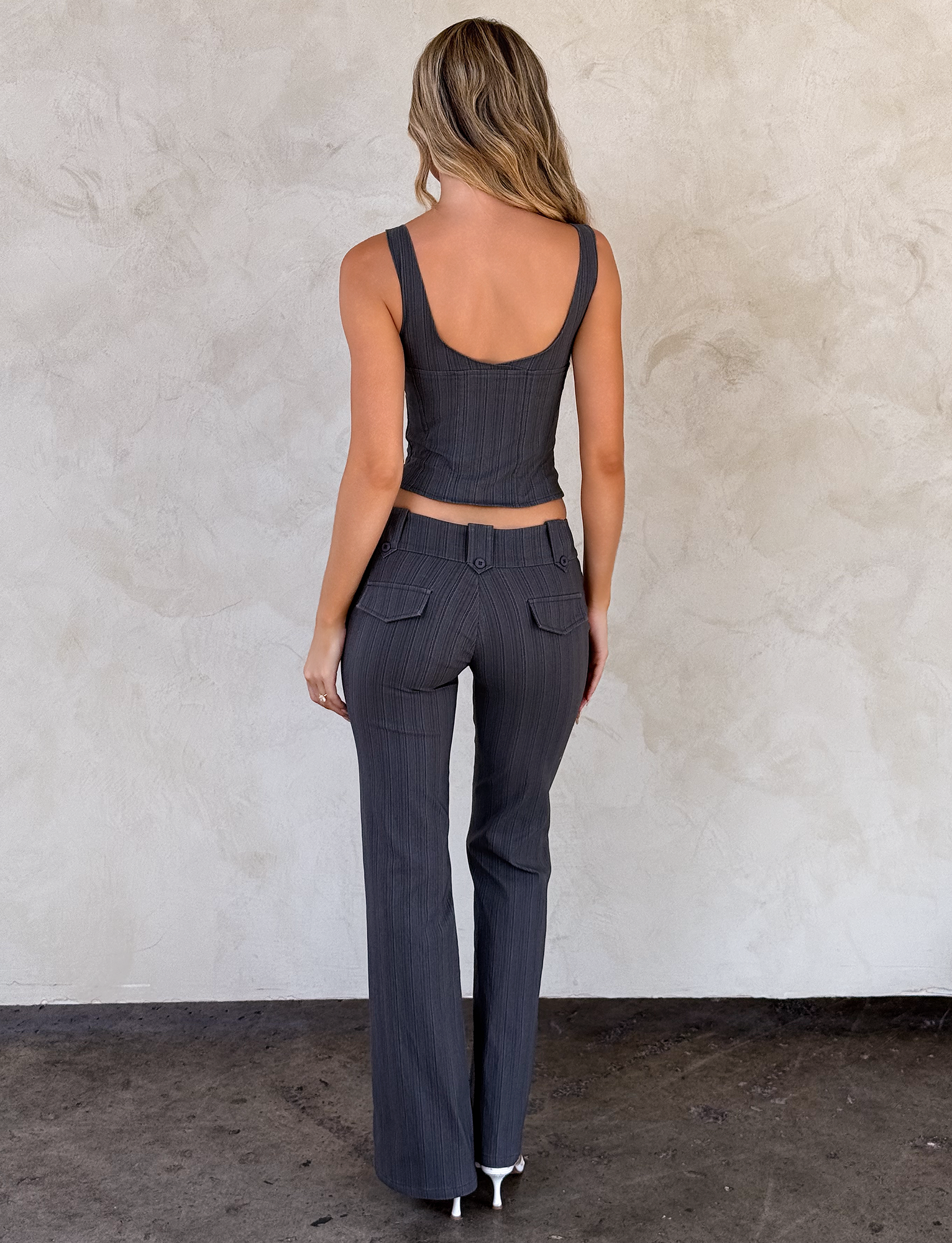 A woman with long wavy hair stands facing a light wall, wearing TIGERMIST’s KITTIE PANT - GREY STRIPE with pockets and a matching sleeveless top, paired with white heels. She is shown from the back.