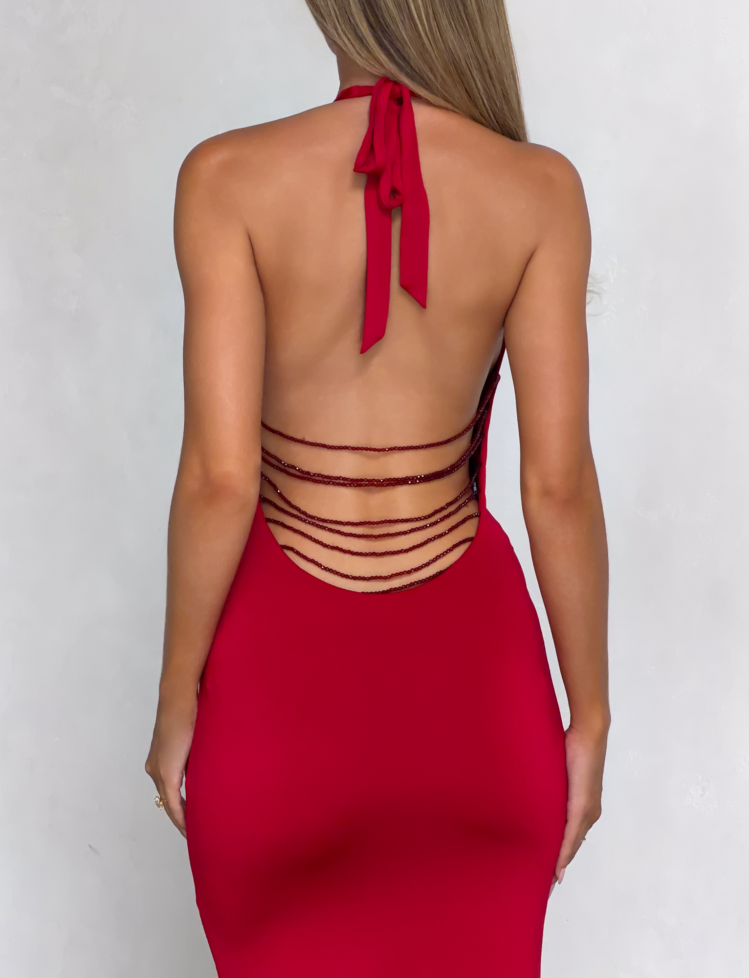A woman stands against a light gray background wearing the TIGERMIST SOLYNE MAXI DRESS in red—a backless halter-neck style with thin horizontal straps across the lower back.