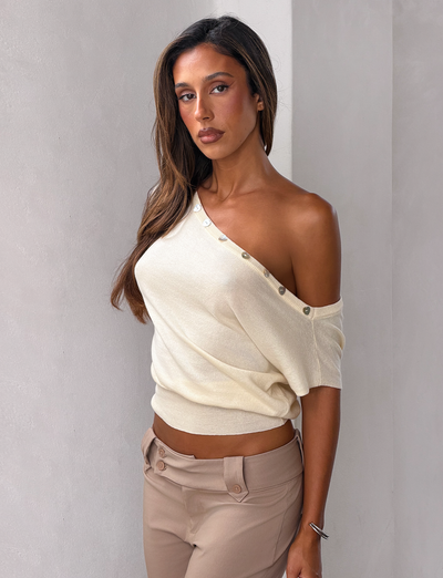A woman with long brown hair wears the TIGERMIST TYLA SLEEVELESS KNIT TOP in cream and beige pants, standing against a light-colored wall and looking at the camera with a neutral expression.