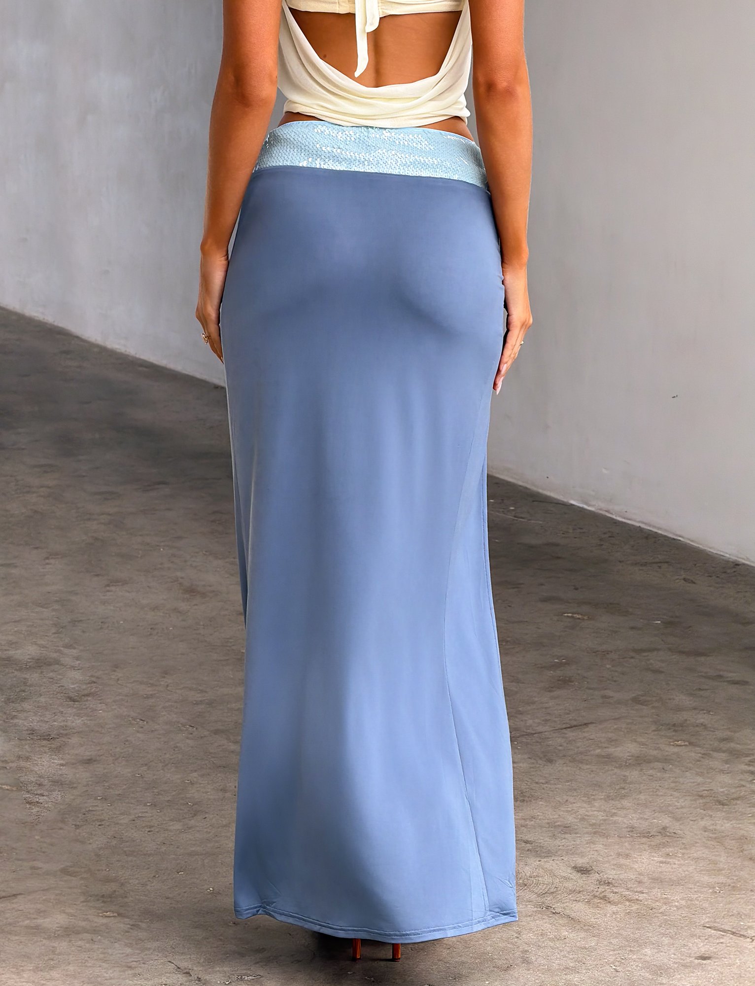A person, shown from the back, wears TIGERMIST's THARA MAXI SKIRT in Slate Blue with a thigh-high slit and a white halter top, standing against a gray wall and concrete floor.