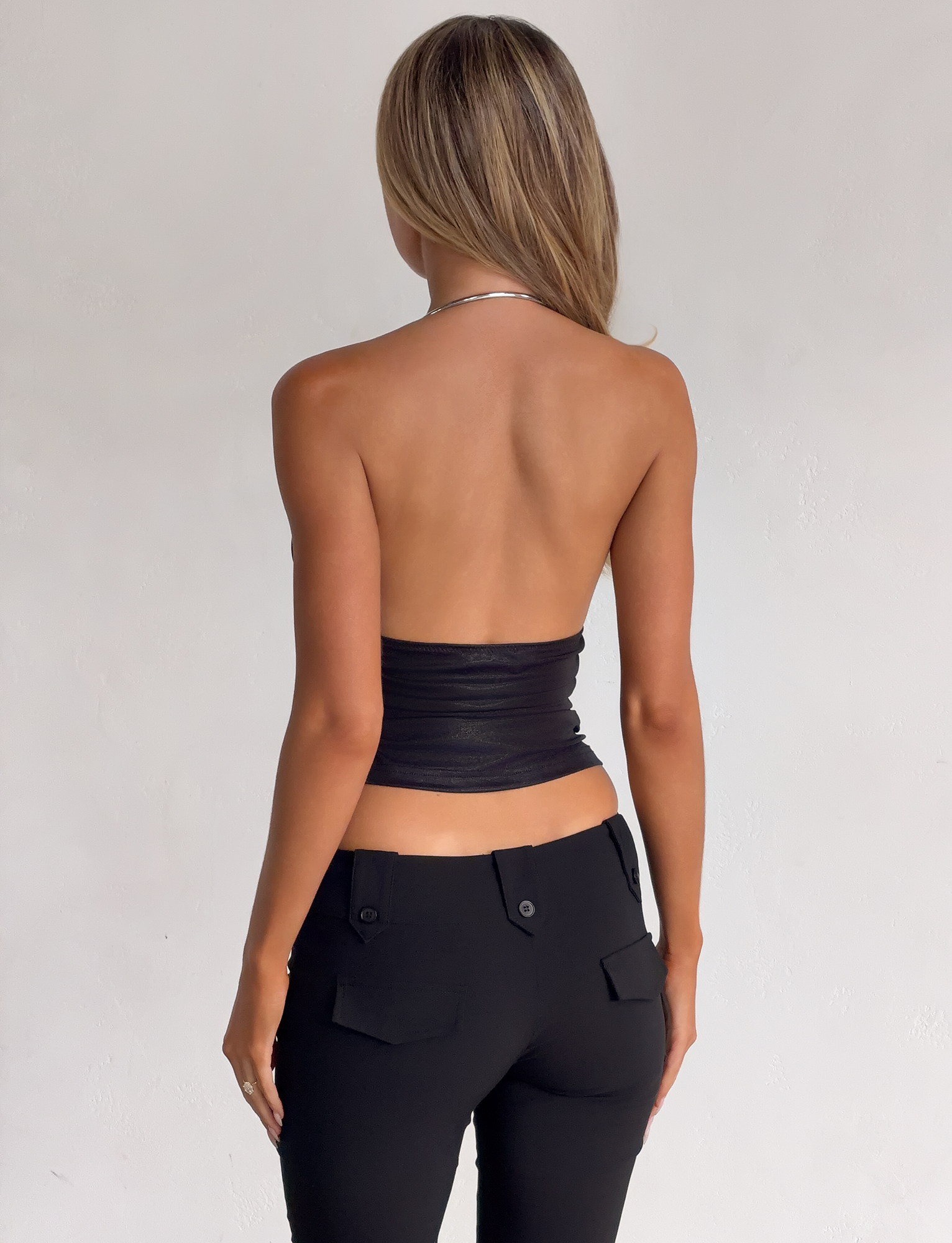 A woman faces away from the camera, wearing the TIGERMIST VIANNA TOP in black metallic and black pants with pockets, set against a plain light background.