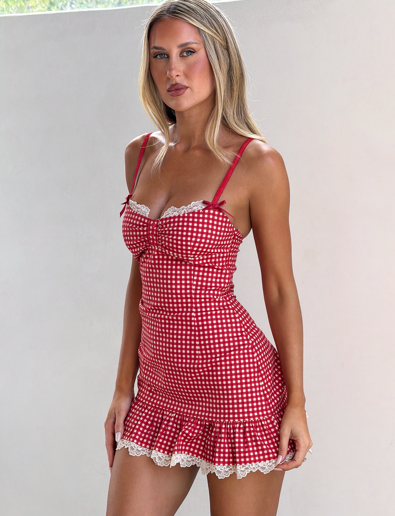A woman with long blonde hair wears the TIGERMIST EMMALINE MINI DRESS in red gingham with lace trim and thin straps, standing against a plain white wall.