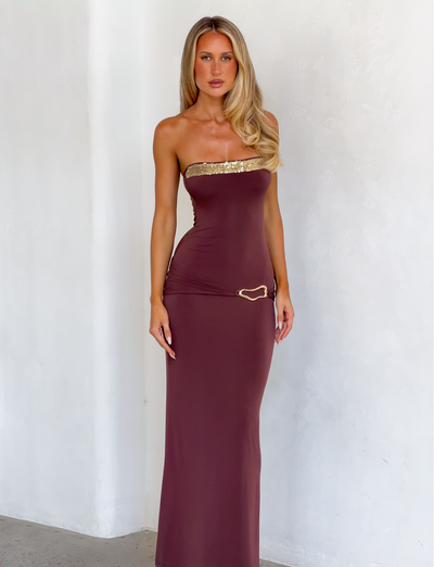 A woman with long blonde hair wears the TIGERMIST EOS SEQUIN TUBE MAXI DRESS in Cinnamon Chocolate, a strapless, body-hugging column silhouette with a gold heart accent at the hip, standing against a plain light-colored wall.