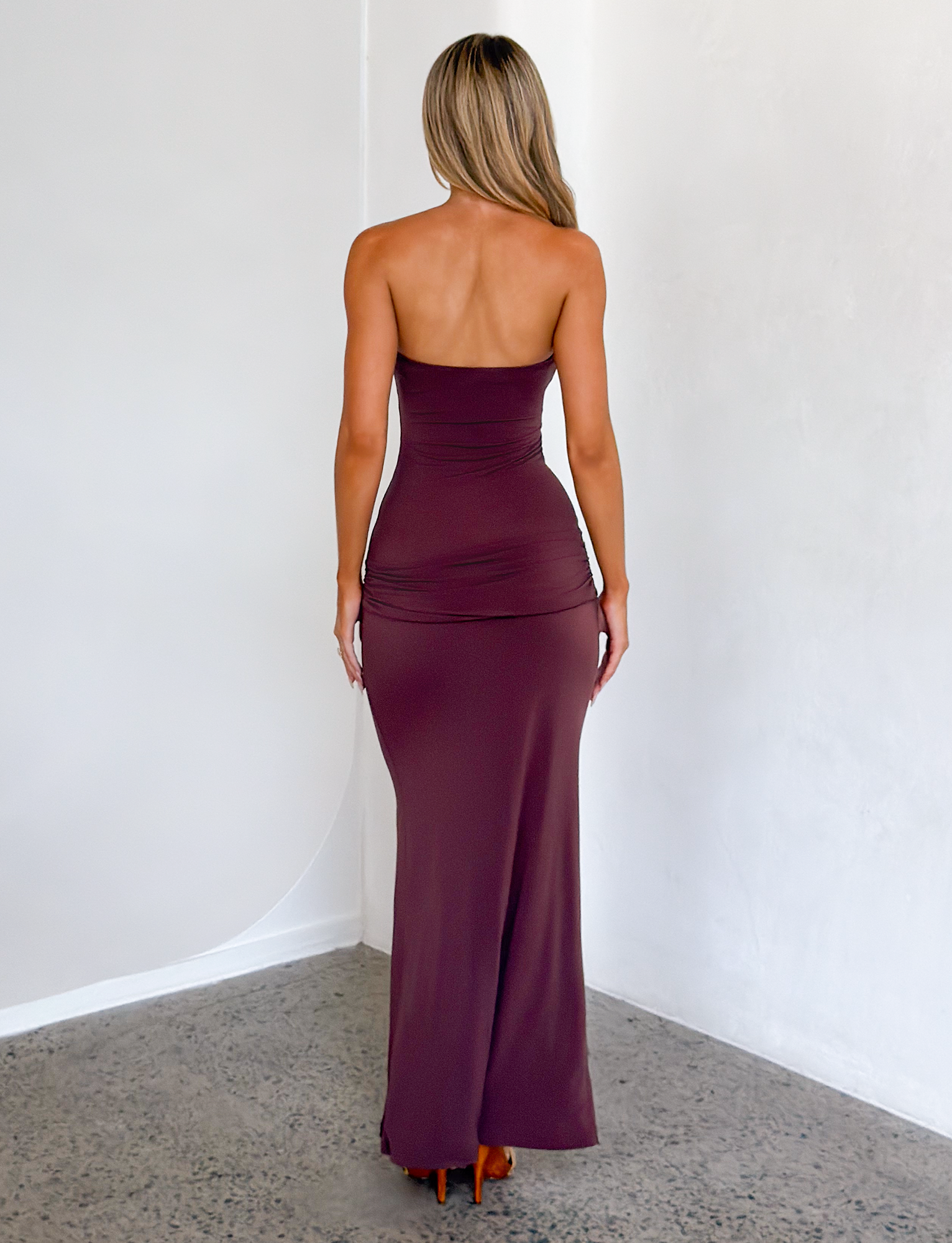 A woman with long blonde hair wears the TIGERMIST EOS SEQUIN TUBE MAXI DRESS in Cinnamon Chocolate, standing with her back to the camera in a minimalist room with light walls and a polished concrete floor.