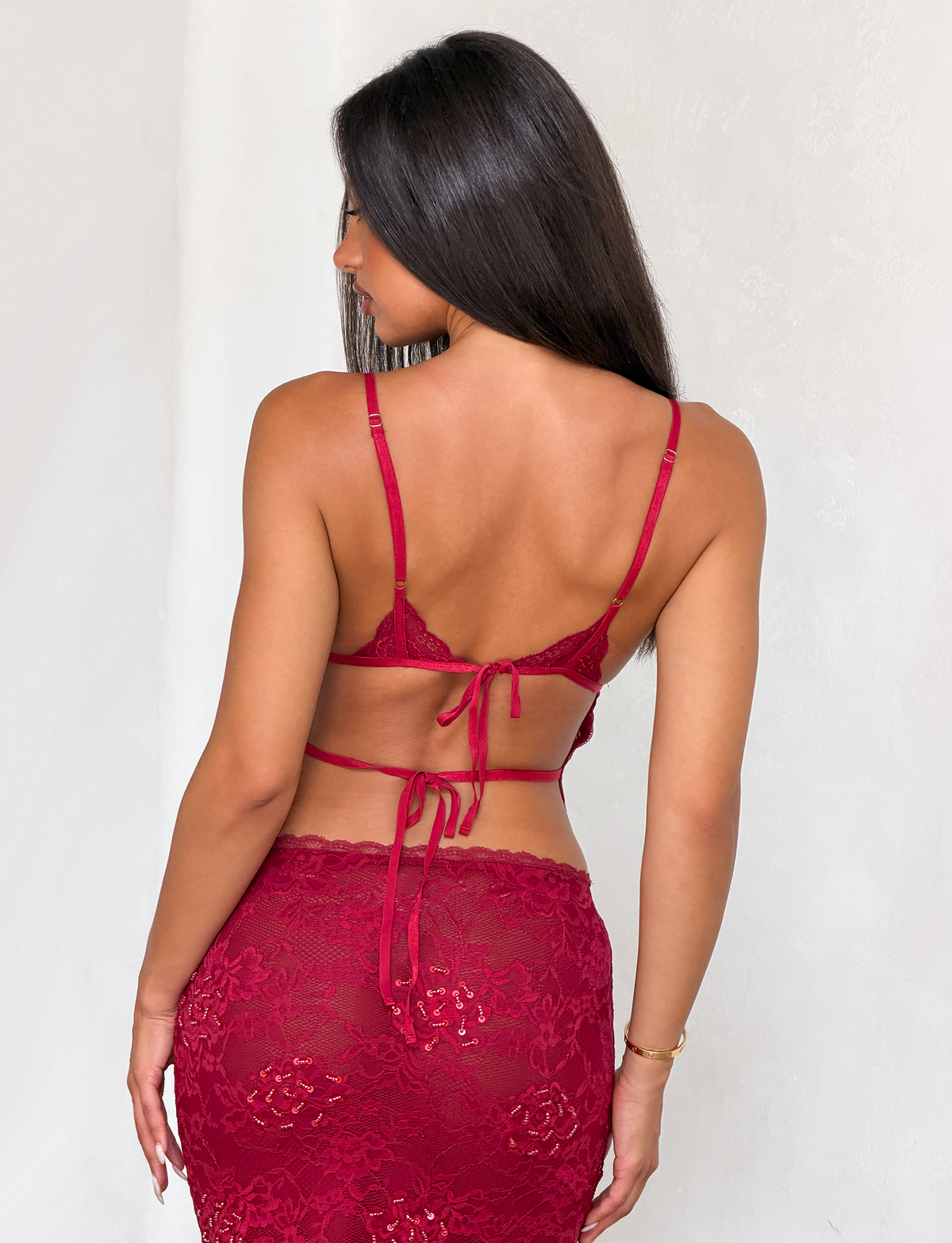 A woman with long dark hair is seen from behind wearing the TIGERMIST KIRRA BEADED SEQUIN LACE TOP in red, paired with a matching high-waisted skirt with a tie-back detail, standing against a light-colored wall.