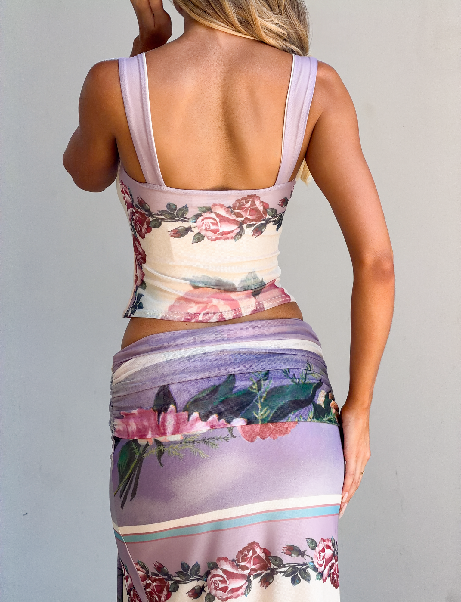 A woman with long blonde hair faces away, wearing the TIGERMIST RAFFIE PRINTED CORSET in Postcard Floral, paired with a matching skirt in purple, pink, and cream. Her arm is bent, set against a plain light background.