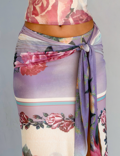 A close-up of a person wearing the TIGERMIST RAFFIE MAXI SKIRT in Postcard Floral, featuring a gathered waist tie and soft roses in pink, red, and purple pastel hues.