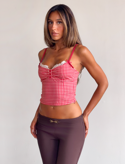 A woman with long, straight hair and a neutral expression stands against a light background wearing the TIGERMIST EMMALINE BUSTIER in red gingham with lace trim and adjustable straps, paired with brown pants. Her arms rest by her sides.