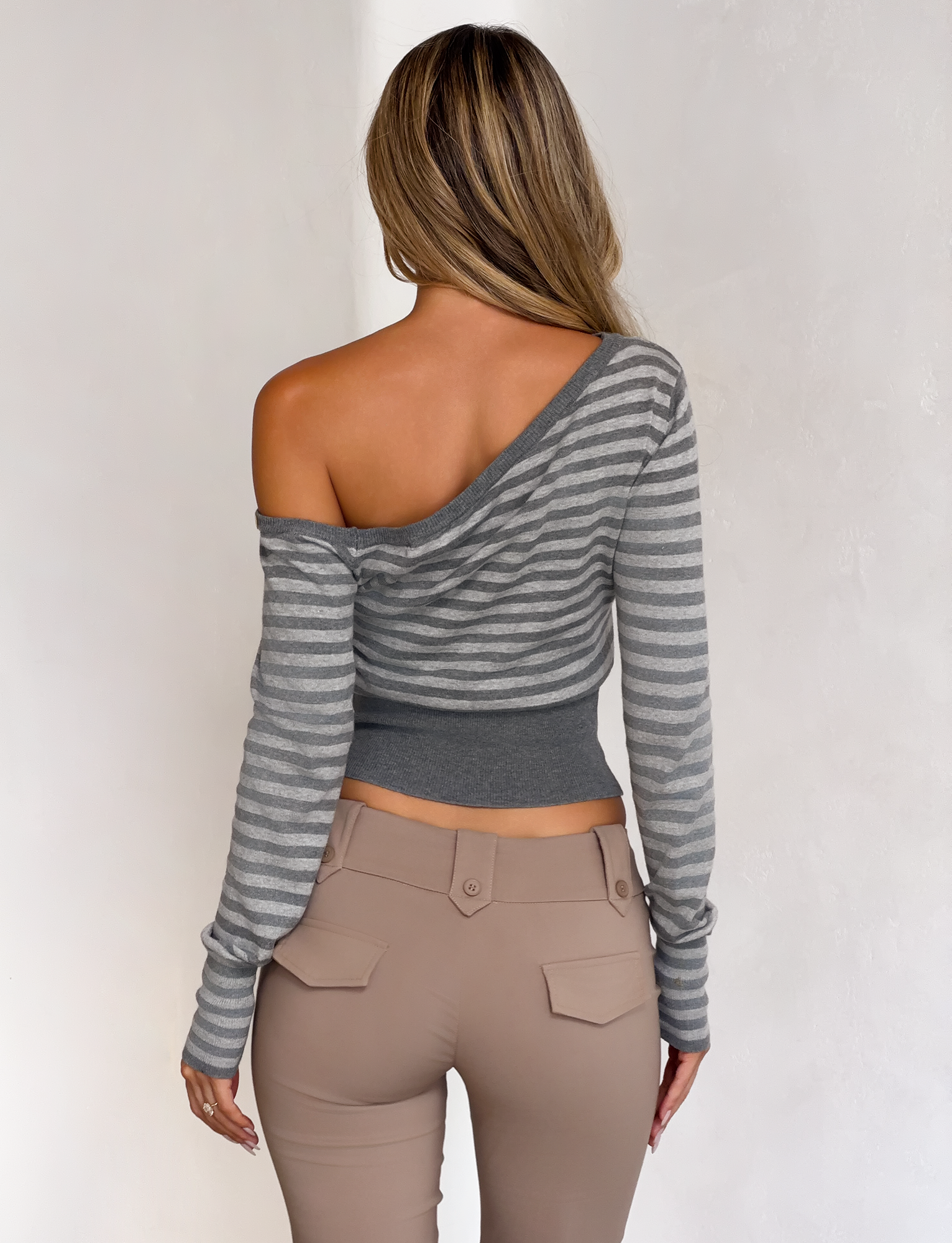 A woman with long, wavy hair stands with her back to the camera wearing the TIGERMIST TYLA JUMPER in grey stripe, styled off-the-shoulder with high-waisted beige pants against a simple light background.