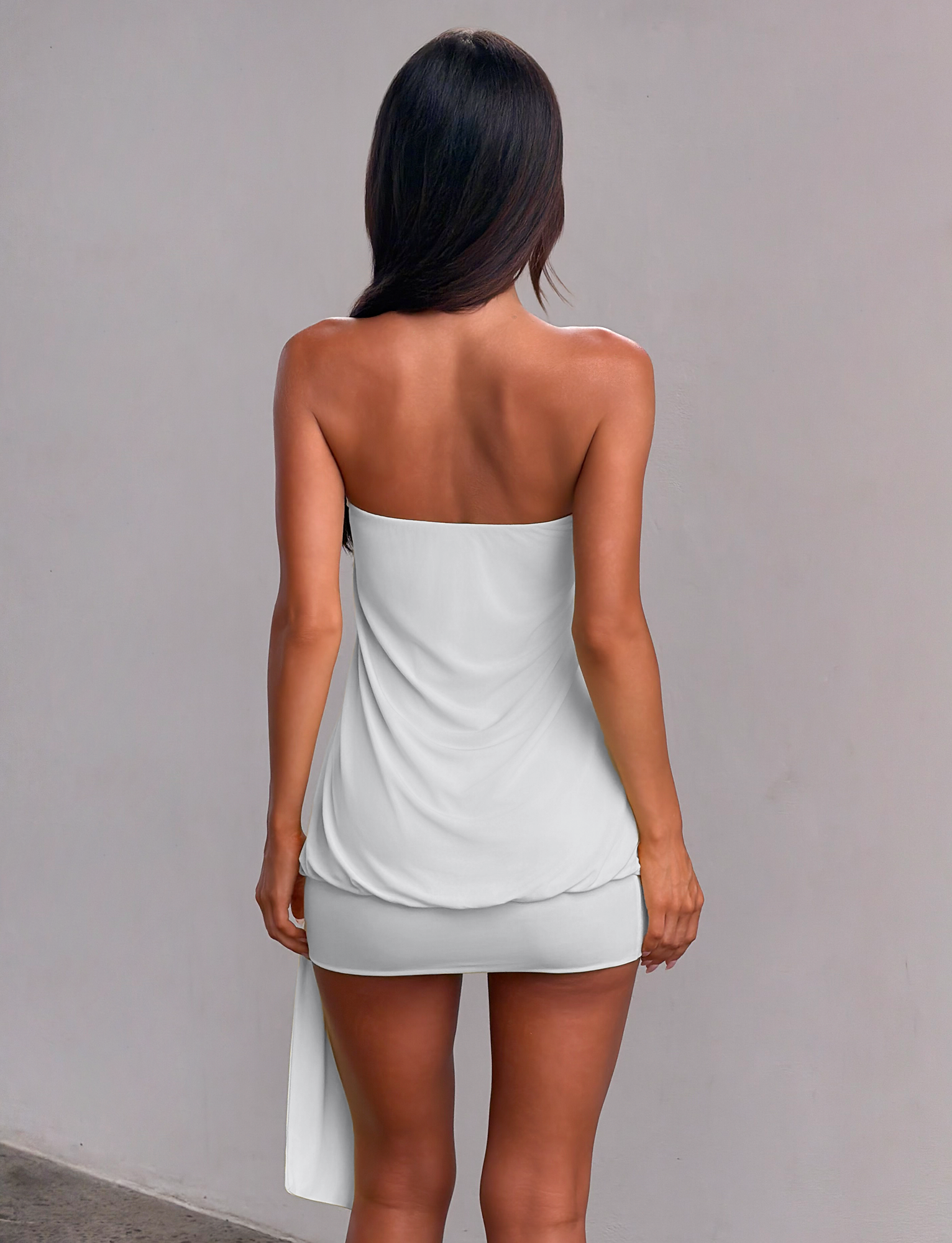 A woman with long dark hair faces a light gray wall, wearing the TIGERMIST RAVENNA MINI DRESS in Cool Slate—a strapless bodycon mini dress with an asymmetrical drop waist skirt that drapes at the back, exposing her shoulders and upper back.