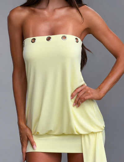 A woman models the TIGERMIST RAVENNA MINI DRESS in butter yellow, a strapless, draped bodycon style with silver eyelet trim along the top, confidently posing with one hand on her hip.