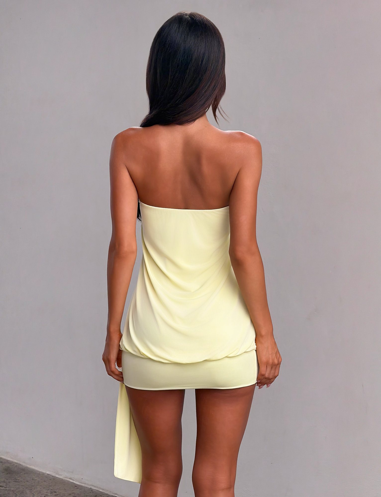 A woman with long dark hair faces away from the camera, wearing the TIGERMIST RAVENNA MINI DRESS in butter yellow, featuring a strapless design and draped fabric detail, set against a plain light gray wall.