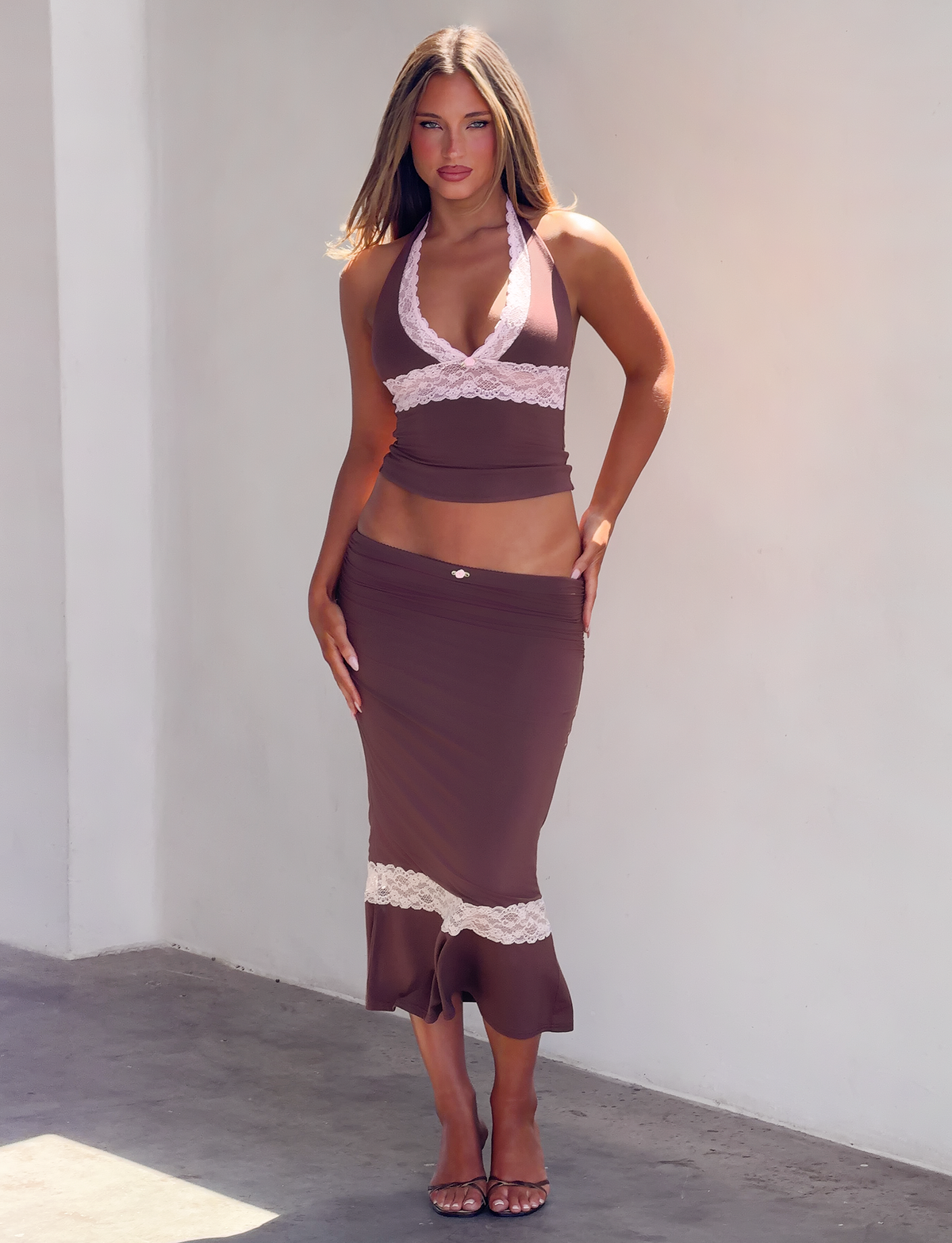 A person wearing a brown halter top with pink lace detail and the matching CALINA LACE PANEL MIDI SKIRT - CHOCOLATE in a mermaid silhouette with a pink lace hem panel.