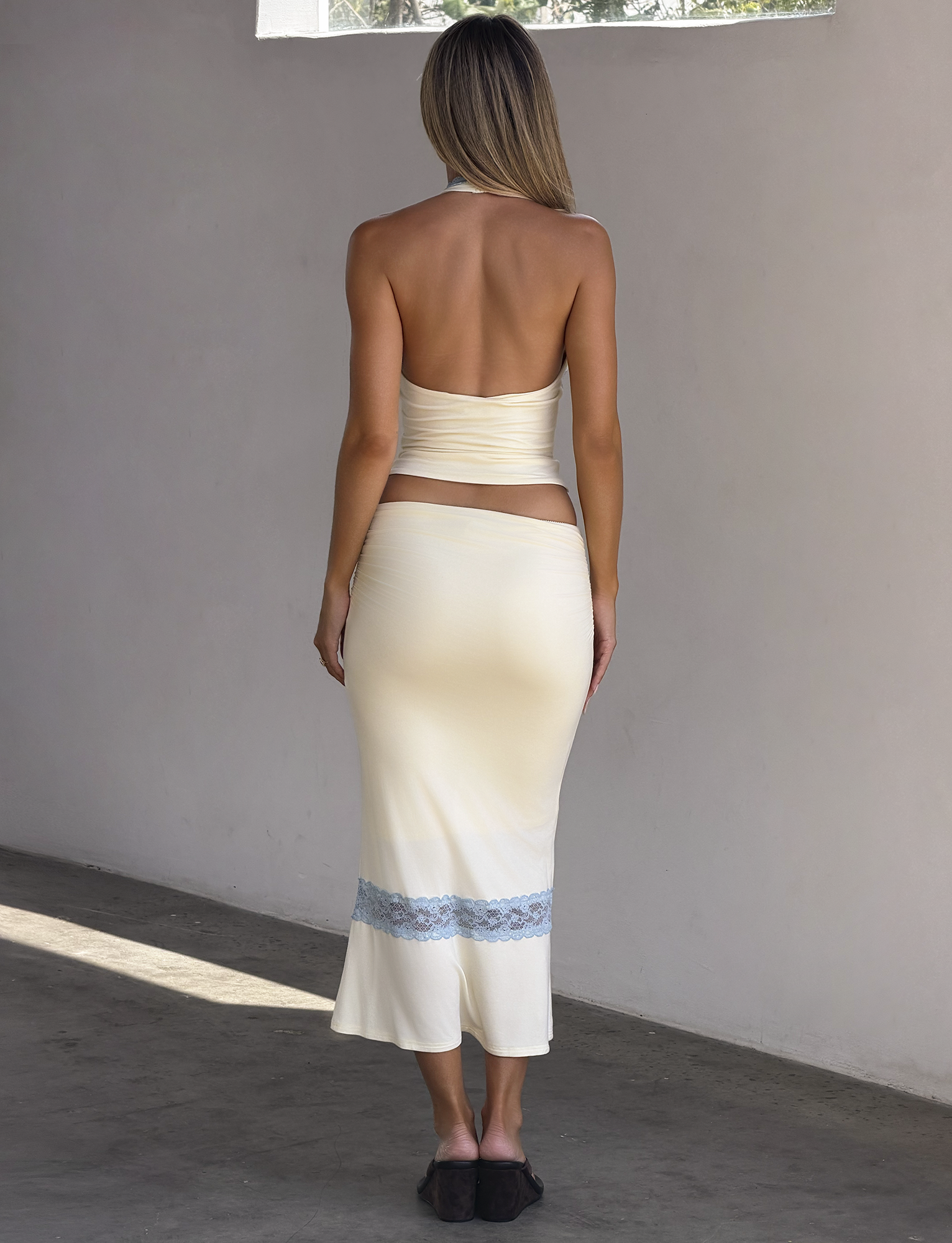 A woman with long, straight hair stands indoors barefoot on a concrete floor, wearing the TIGERMIST CALINA LACE PANEL MIDI SKIRT in cream, featuring blue lace details at the hem and paired with a backless halter top.