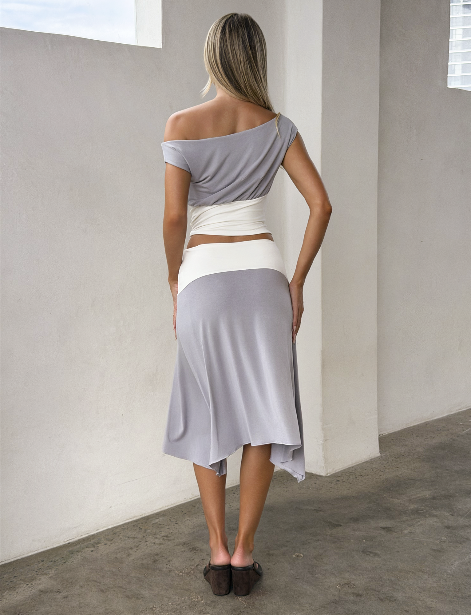 A woman indoors, facing away, wears the TIGERMIST ALYSSA HI LO SKIRT in grey with an asymmetrical hem and front slit, paired with a light gray off-shoulder top, white waist wrap, and brown sandals.