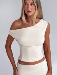 A woman with long blonde hair wears the TIGERMIST ALYSSA OFF SHOULDER TOP in cream, featuring side buttons, paired with a matching skirt, standing against a plain light background.